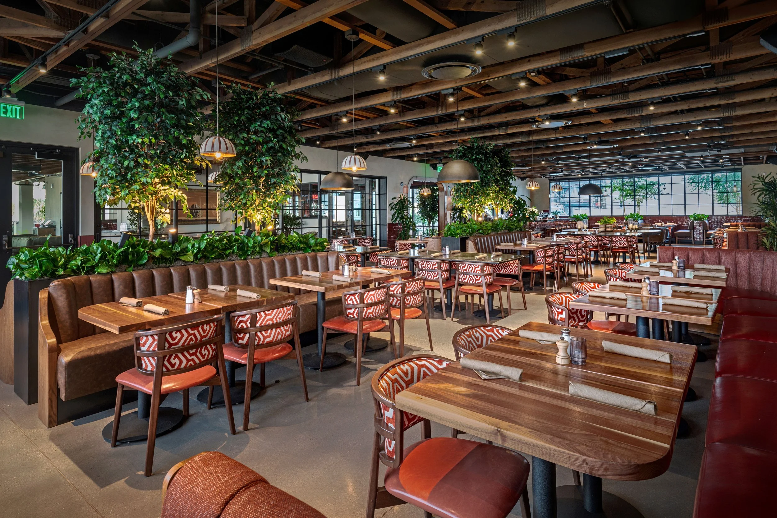 Interior of a modern restaurant with wooden tables and patterned chairs, greenery decor, large windows, and ambient lighting.