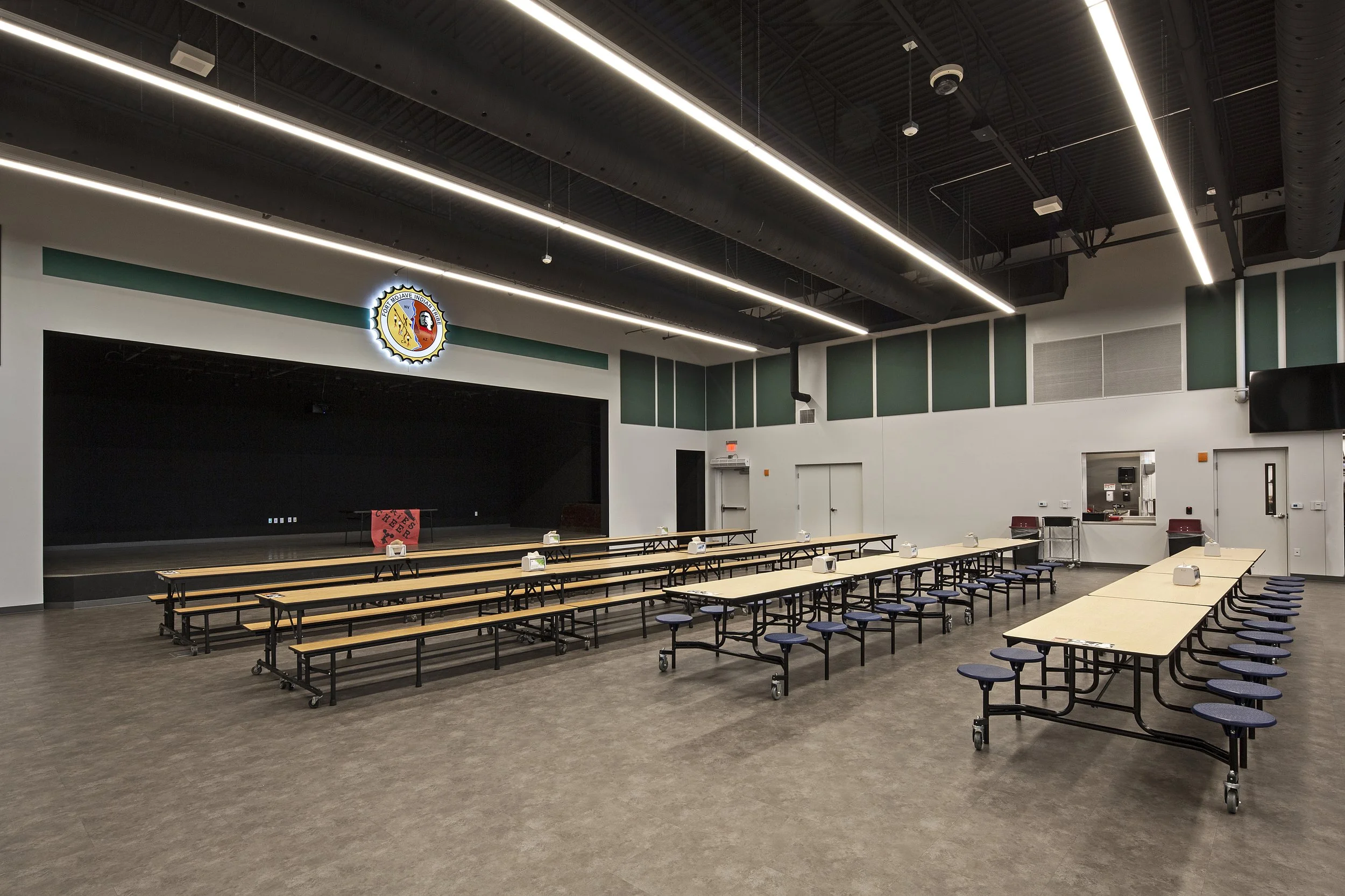 Large community hall with long tables and chairs arranged for a gathering or event. There is a stage at the front with a sign that says "CAMP STAFF BEER." The room has a high ceiling with modern lighting, and a logo on the wall above the stage. Tissu
