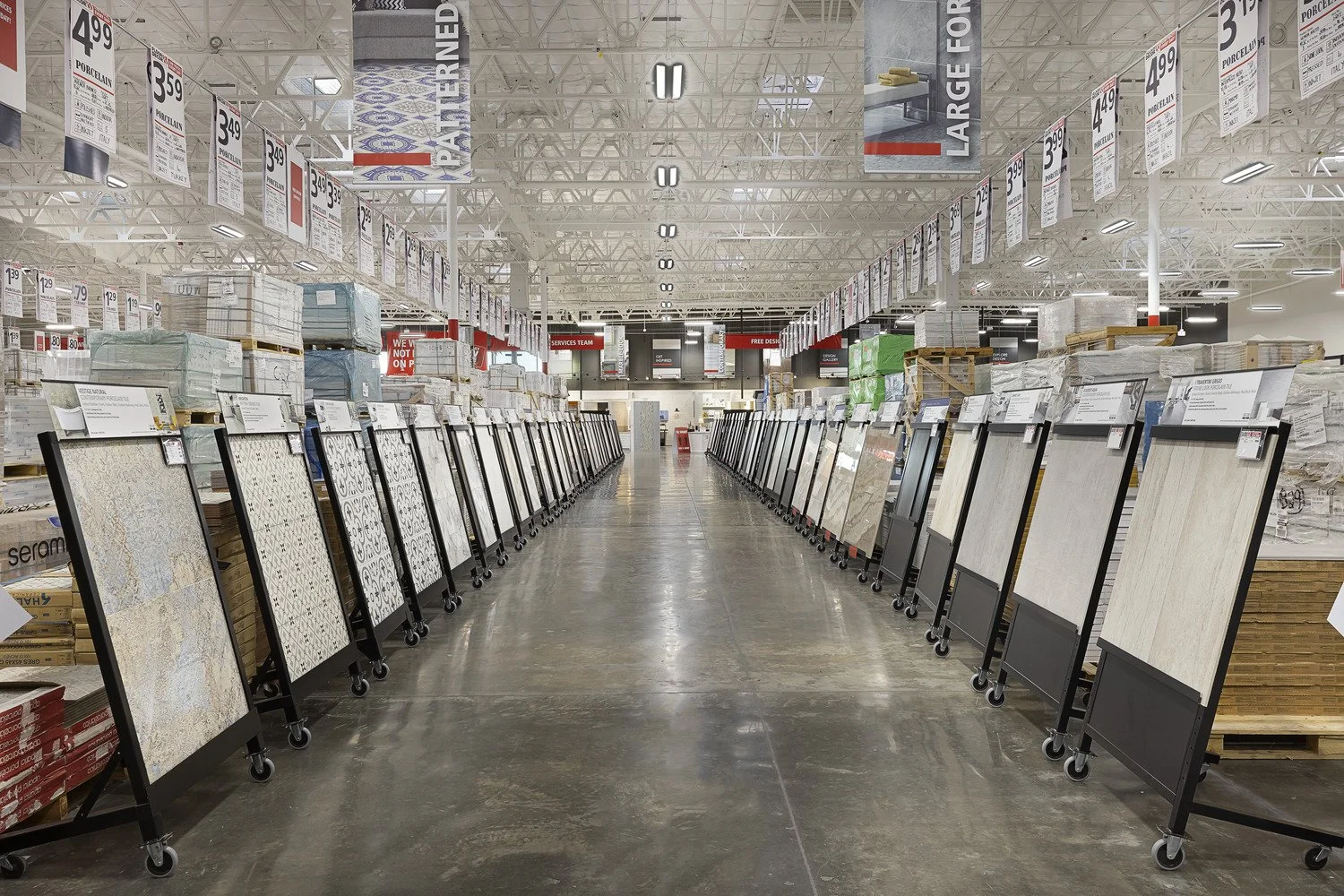 A large home improvement store aisle displaying various tile samples on black mobile stands, with price tags hanging from the ceiling, and stacked boxes of tiles on shelves in the background.
