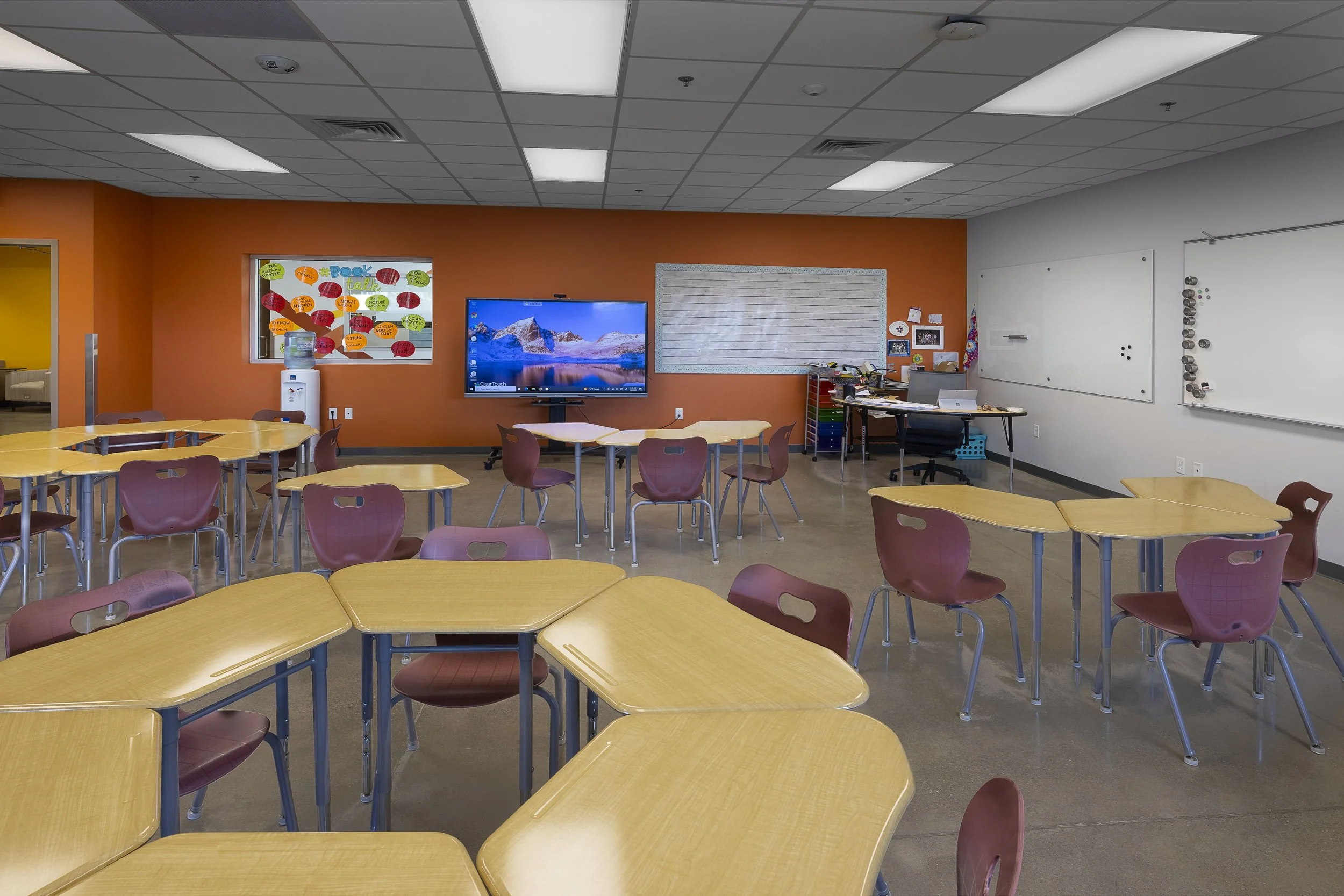 Empty classroom with yellow-topped desks and purple chairs, white and orange walls, a whiteboard, a computer desk, a large display monitor, and colorful posters.