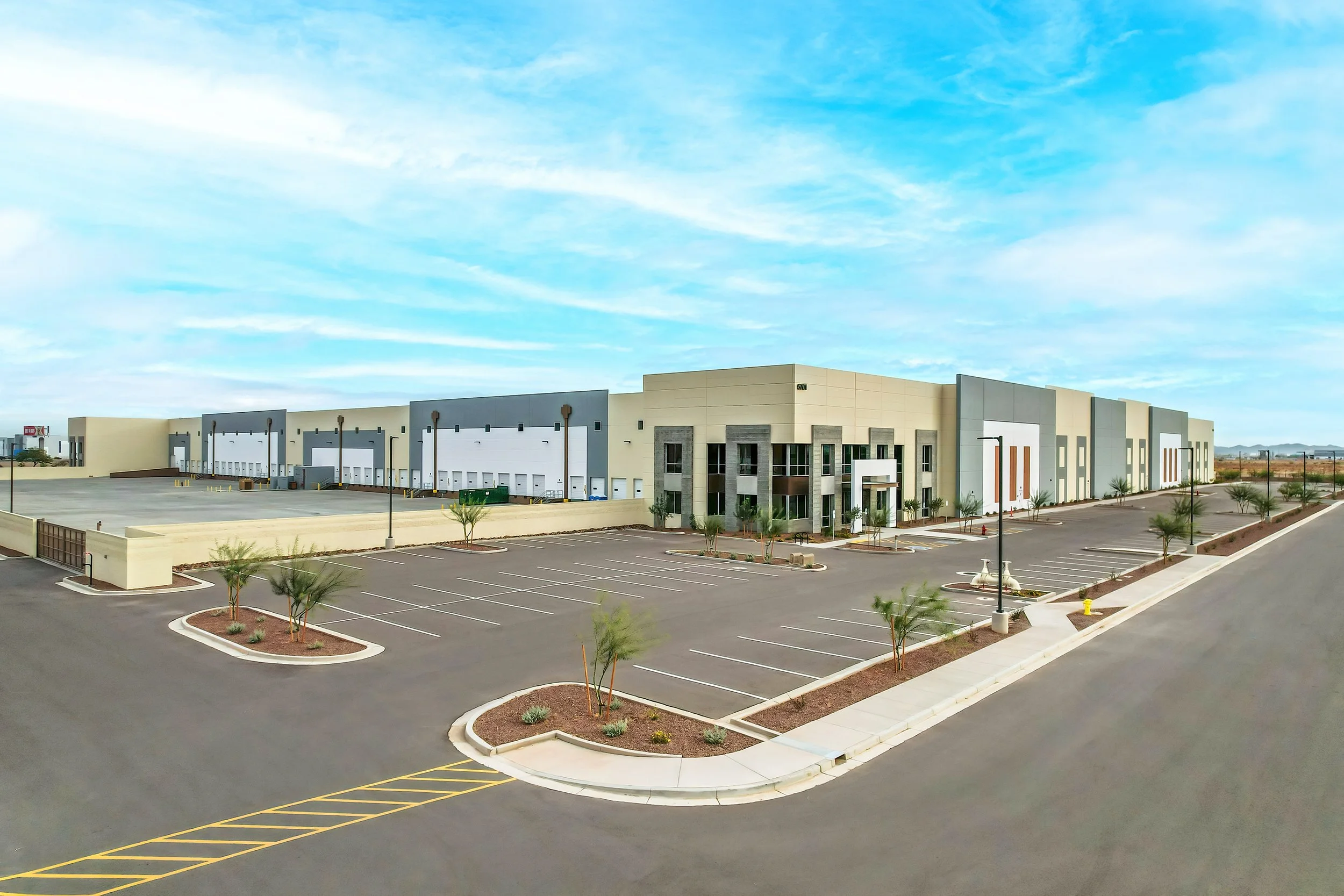 Large modern warehouse with a spacious parking lot and small trees along the sidewalk, under a blue sky with wispy clouds.