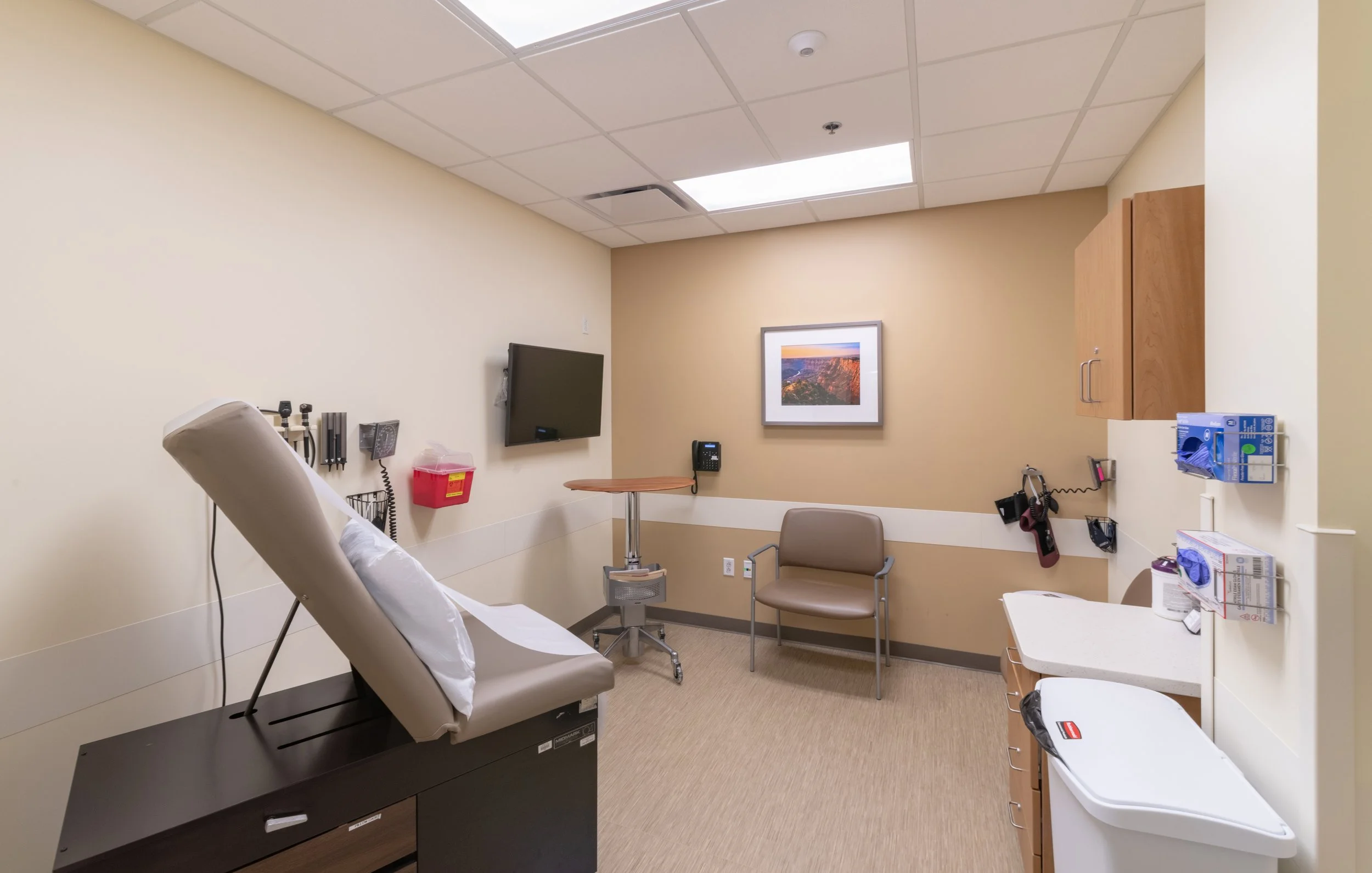 Interior of a medical examination room with a patient bed, wall-mounted television, framed picture, chairs, medical supplies, and a countertop.