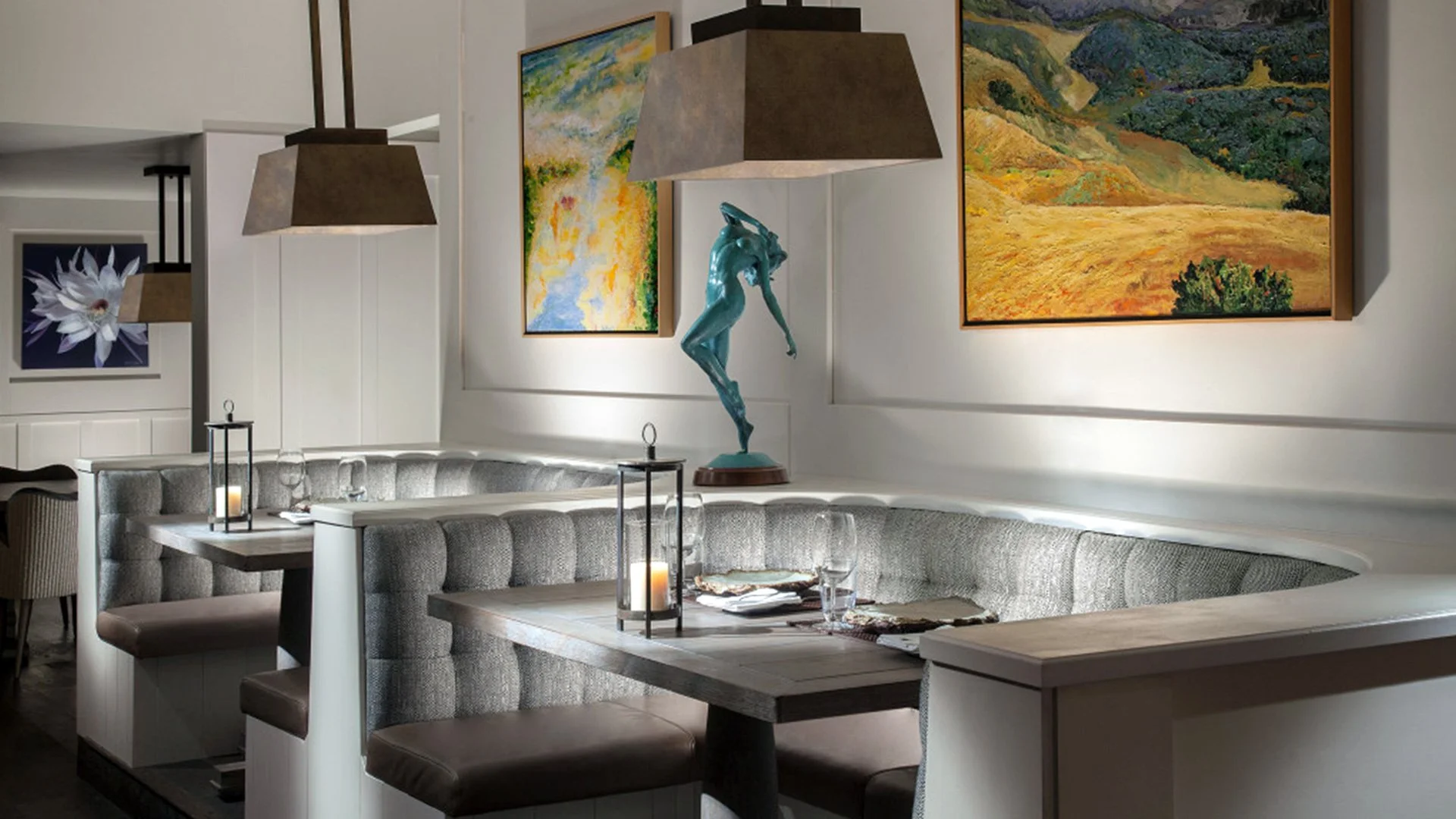 Interior of a restaurant with a curved booth, hanging pendant lights, colorful landscape paintings, a blue sculpture of a woman, and dining tables set with candles, glasses, and napkins.