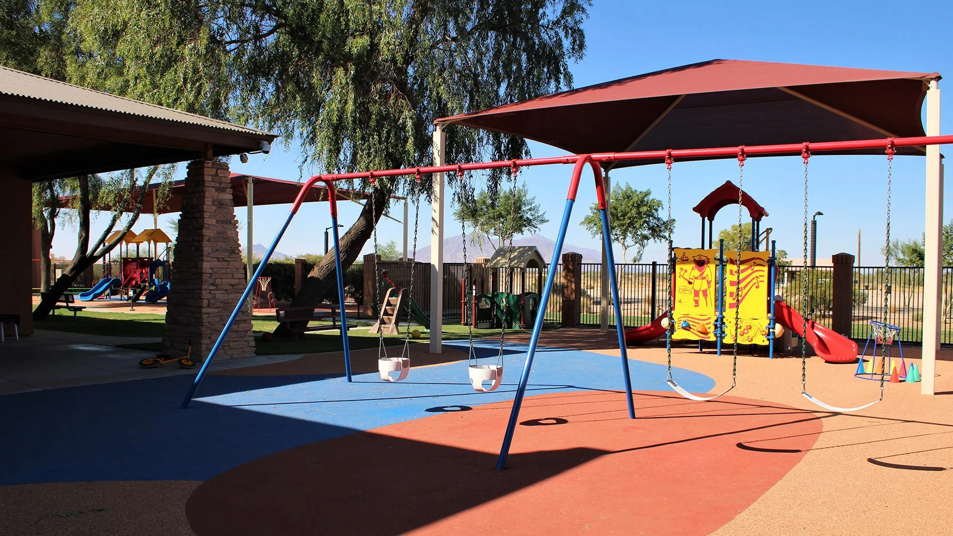 Colorful playground with swings, climbing structures, slides, and cone traffic markers, surrounded by trees and a fence.
