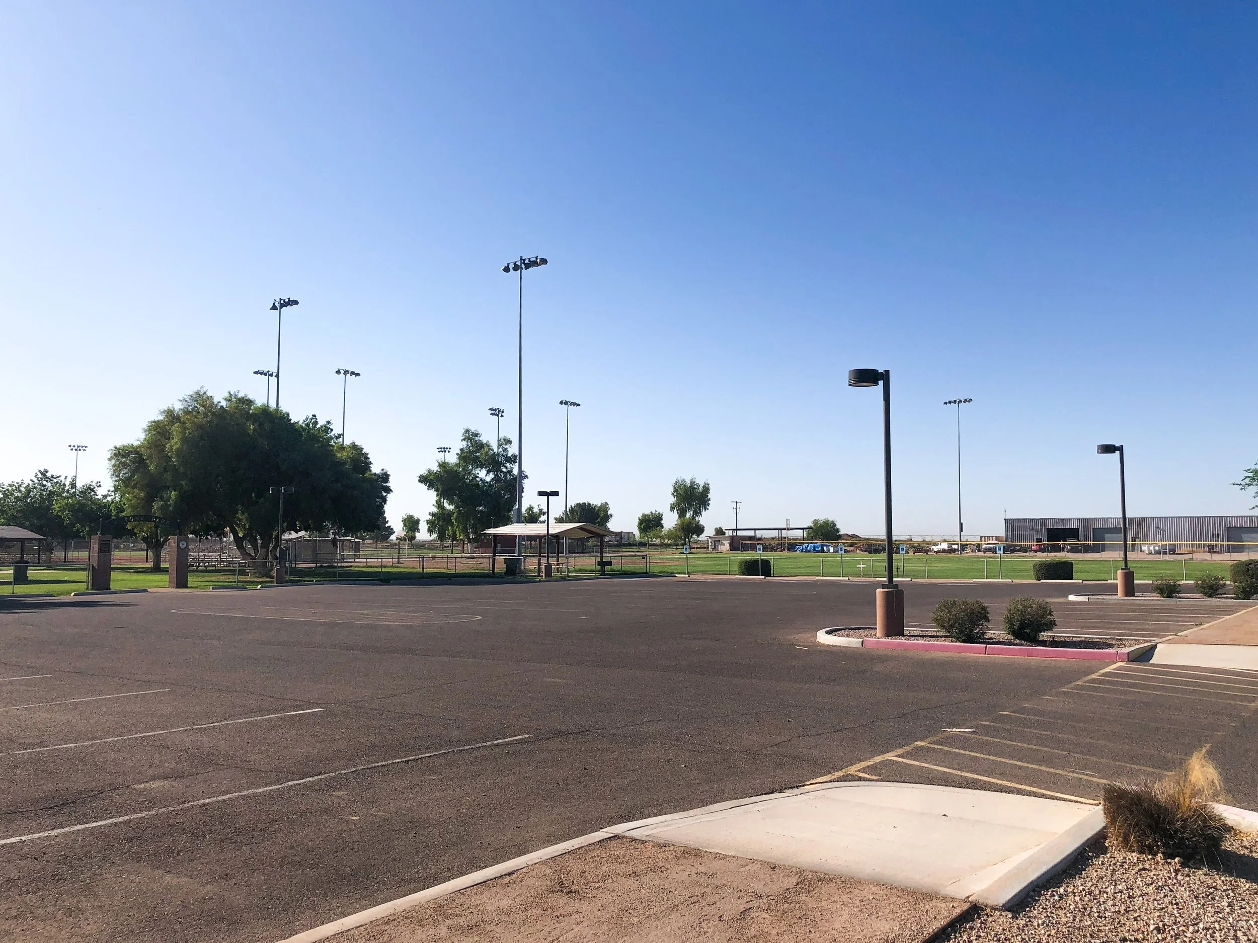 Empty parking lot with trees, lamp posts, and a sports field in the background under a clear blue sky.