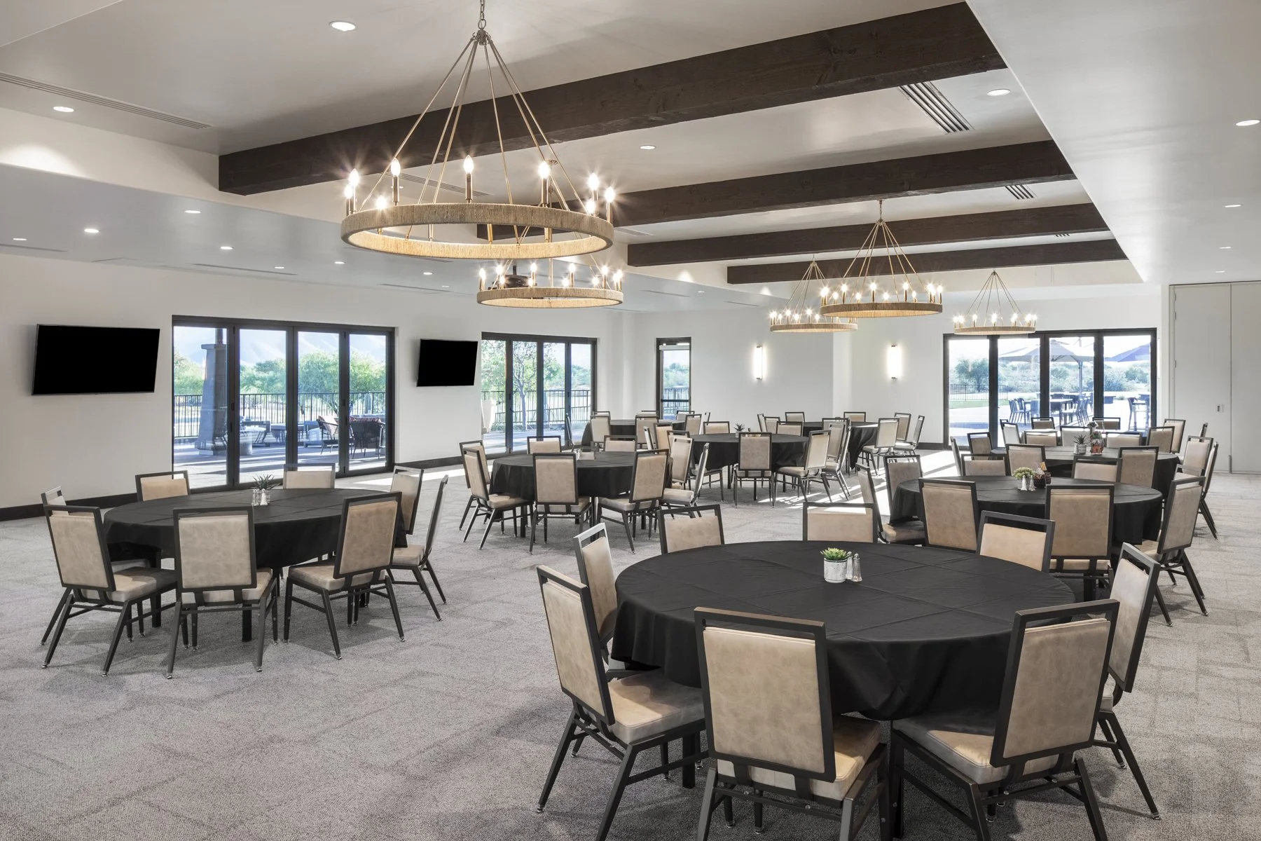 A spacious banquet hall with round tables covered in black tablecloths and beige chairs. Large windows and sliding glass doors let in natural light, revealing an outdoor patio area. Elegant chandeliers hang from the ceiling, and wall-mounted televisi