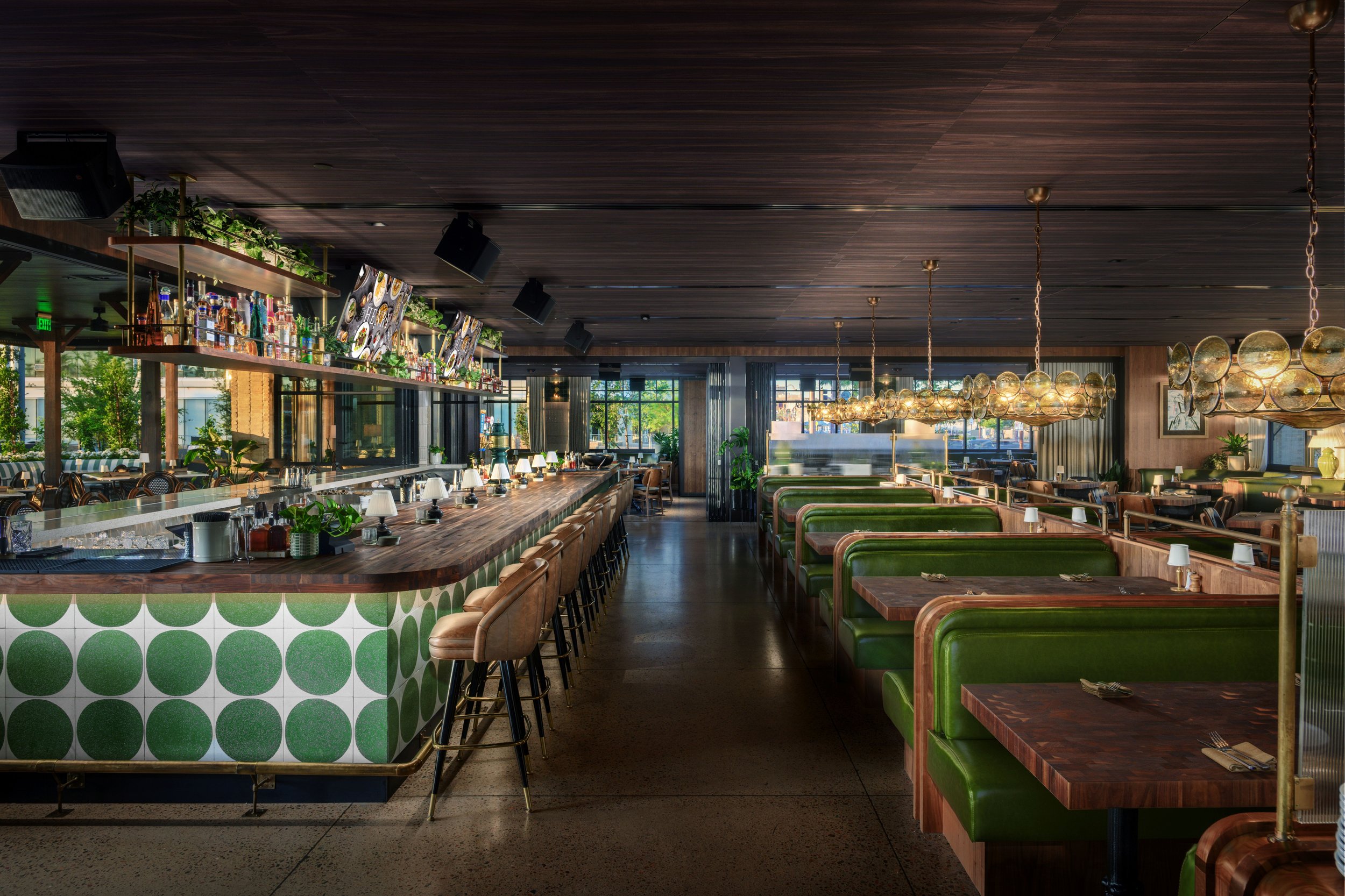 Interior of a modern restaurant with a bar area featuring a curved wooden counter with green circle patterned front, bar stools with beige seats, and booth seating with green cushions and wooden tables. The restaurant has a dark wood ceiling, hanging