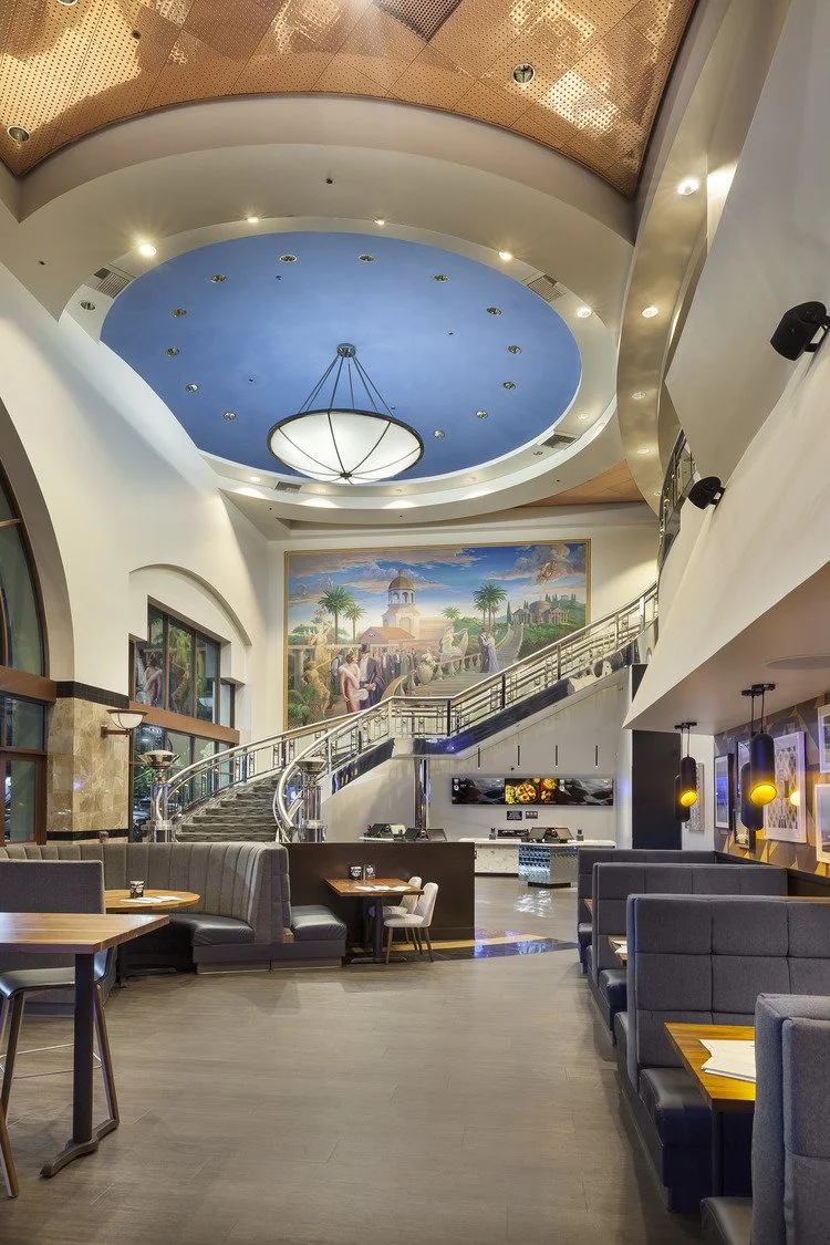 Interior of a modern restaurant or lounge with a high ceiling, a large blue circular ceiling feature with a chandelier, a mural of a historical or scenic scene, and seating booths and tables.