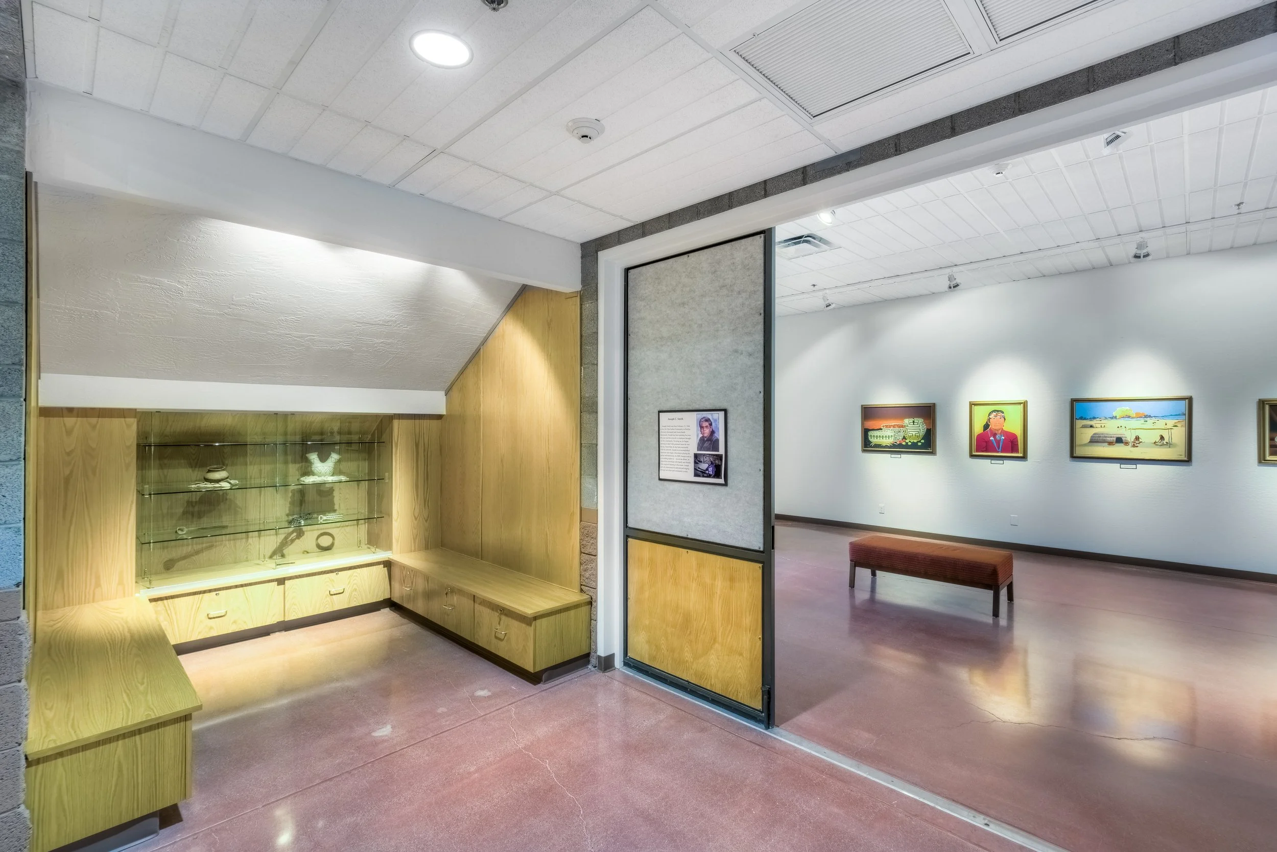 Interior view of an art gallery with framed paintings on the wall and a bench in the center. A corridor on the left side shows a display case with artifacts and display shelves.