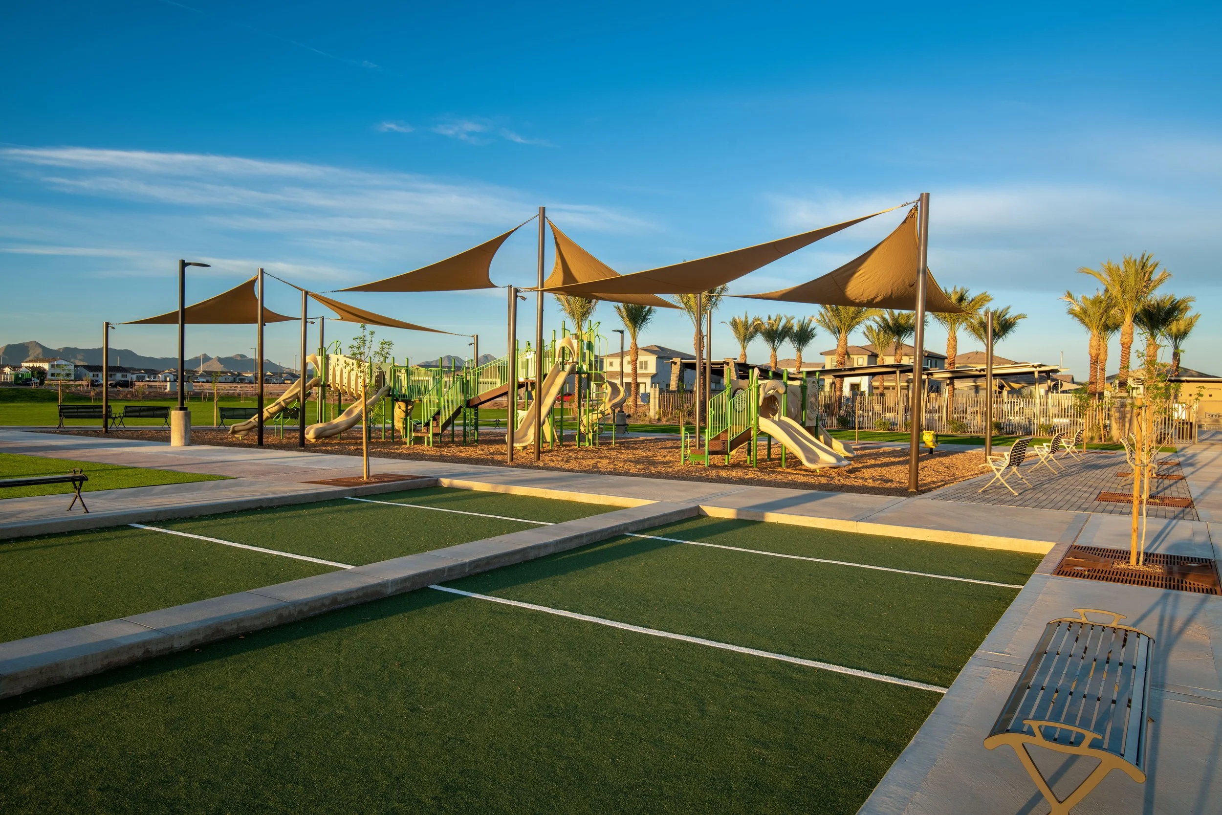 Outdoor playground with slides and climbing structures, shaded by tan sails, surrounded by palm trees, benches, and parking lot in the background, under a clear blue sky.