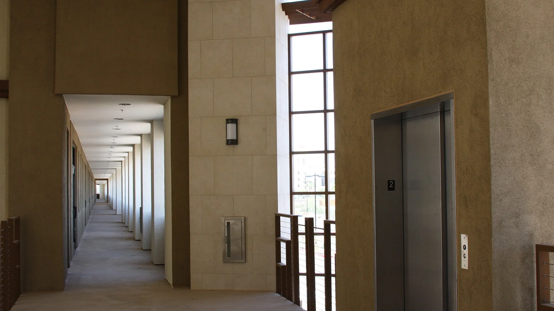 The image shows an indoor corridor with an elevator on the right and a large window revealing a city view in the background. The corridor extends into the distance with doors on the left side.