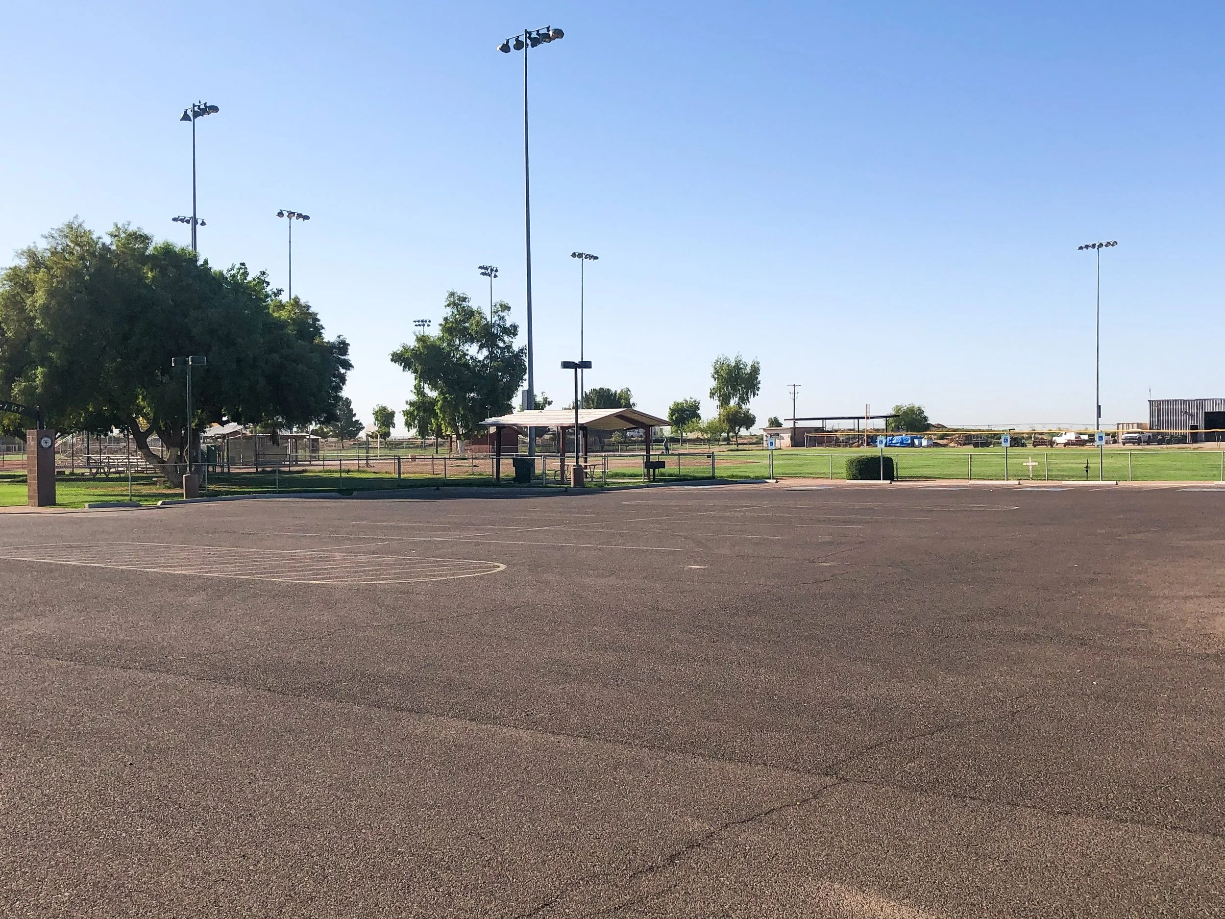 Empty parking lot with sports field and athletic stadium lights in background, some trees and a small shelter in the distance under a clear blue sky.