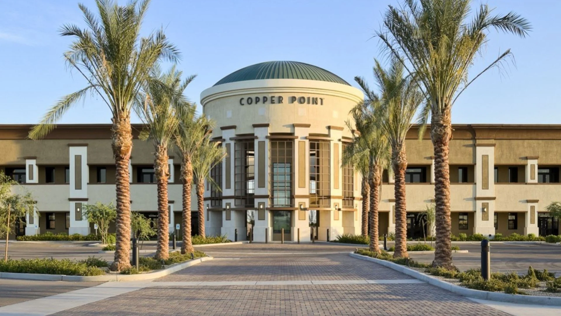 A modern building with a rounded roof labeled 'Copper Point,' surrounded by palm trees and a paved parking lot.