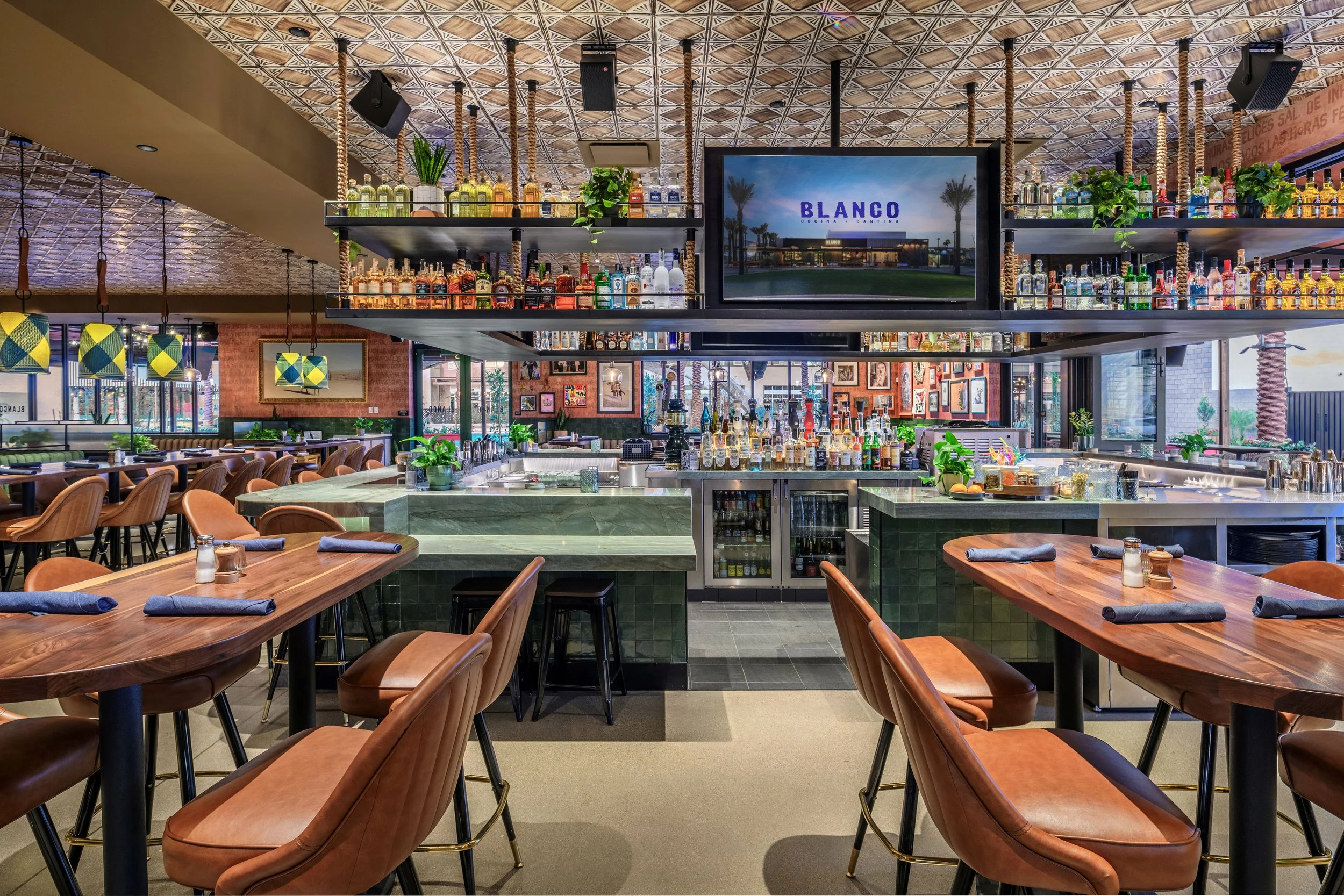Interior of a modern restaurant and bar with wooden tables, leather chairs, and a well-stocked bar area with bottles and plants, featuring a large screen displaying 'Blanco' and palm trees.