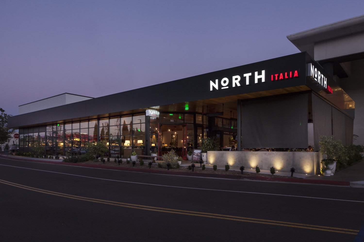 Exterior view of a modern restaurant named 'NORTH ITALIA' with illuminated signage and large glass windows showing the interior, taken during dusk.