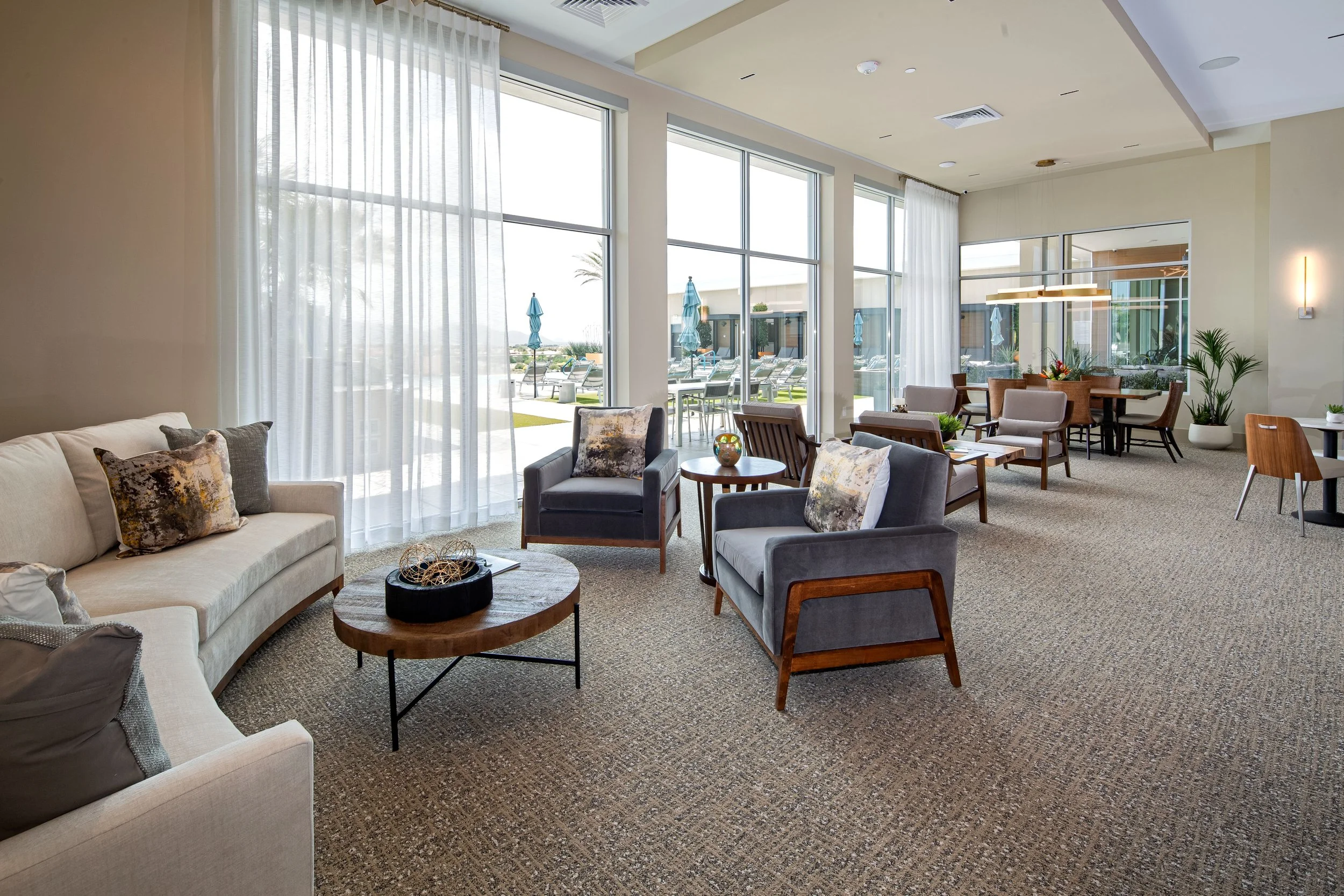 A spacious hotel lounge with large floor-to-ceiling windows and sheer white curtains, furnished with a beige sofa and multiple armchairs, small round tables, and potted plants, with a view of outdoor pool chairs and umbrellas.