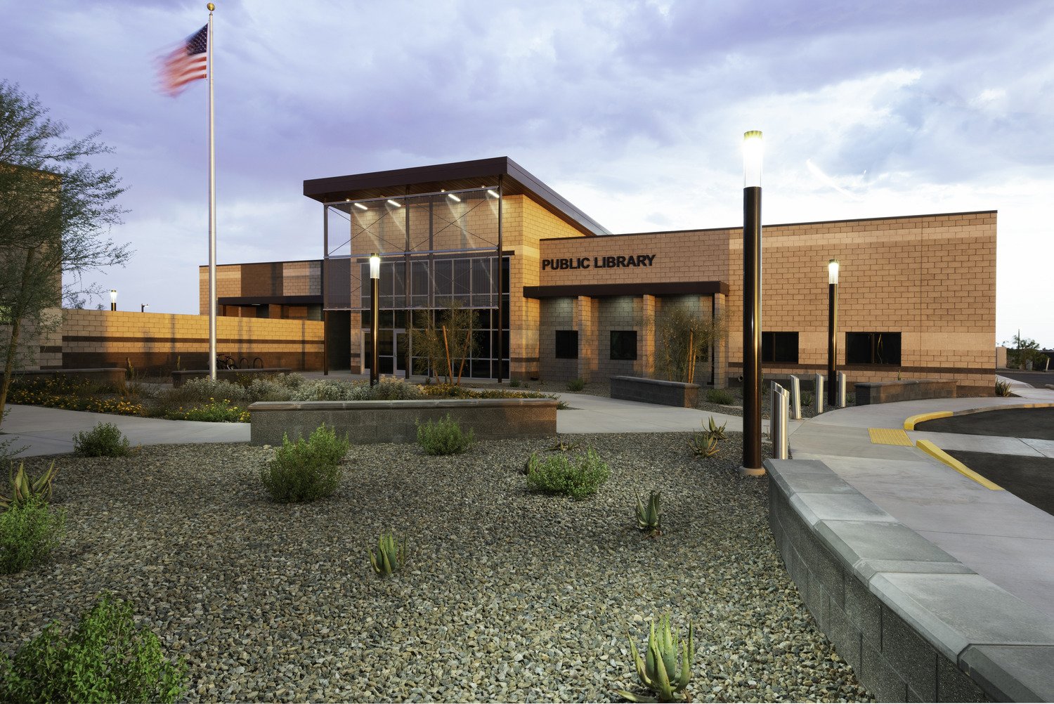 Modern public library building with a sign reading 'Public Library', outdoor landscaping with rocks, small plants, trees, sidewalk, and street lamps, and an American flag flying on a pole outside
