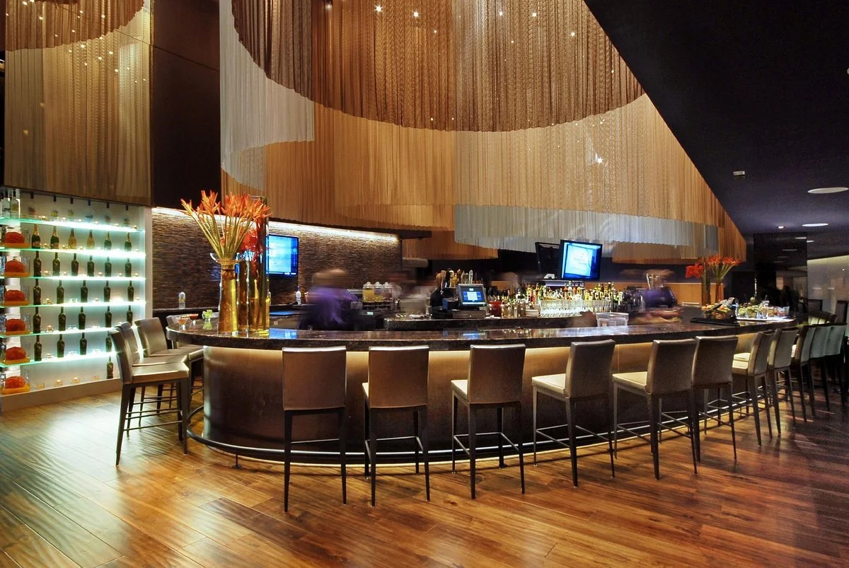 Modern bar with a curved black marble counter, beige chairs, decorative vases with orange flowers, and a backlit display of bottles on the left side, in a softly lit lounge area with wooden floors and an artistic ceiling installation.
