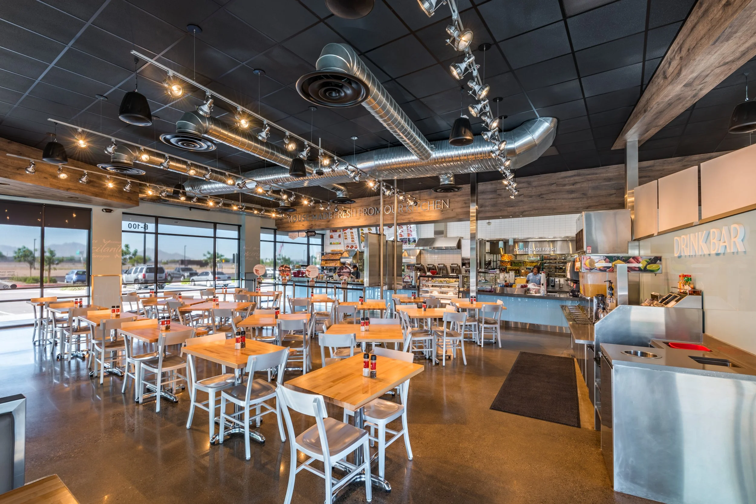Interior of a modern casual dining restaurant with wooden tables and white chairs, large windows, and an industrial-style ceiling with exposed ductwork.
