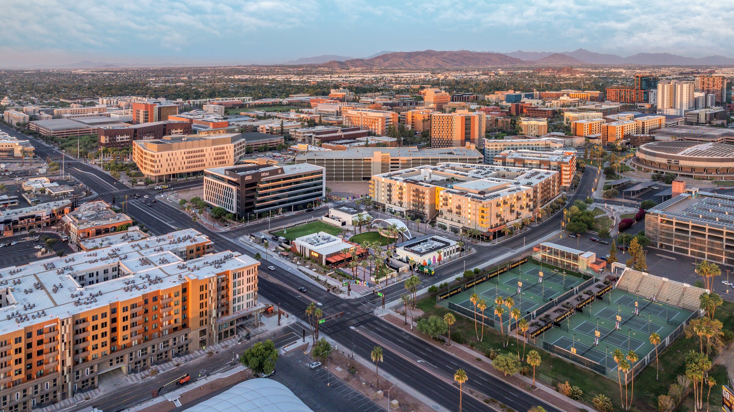 Aerial view of a cityscape featuring modern buildings, roads, tennis courts, and mountains in the background during sunset.