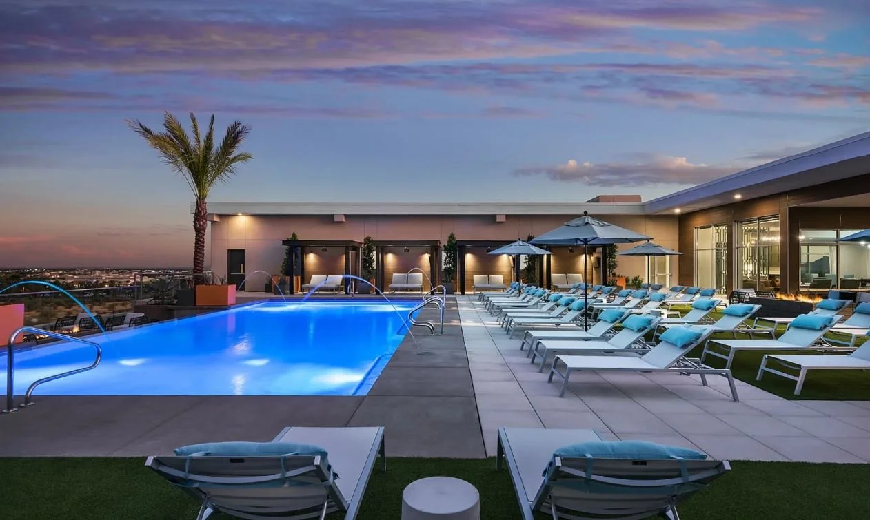Rooftop swimming pool area with lounge chairs and umbrellas at sunset, city view in the background.