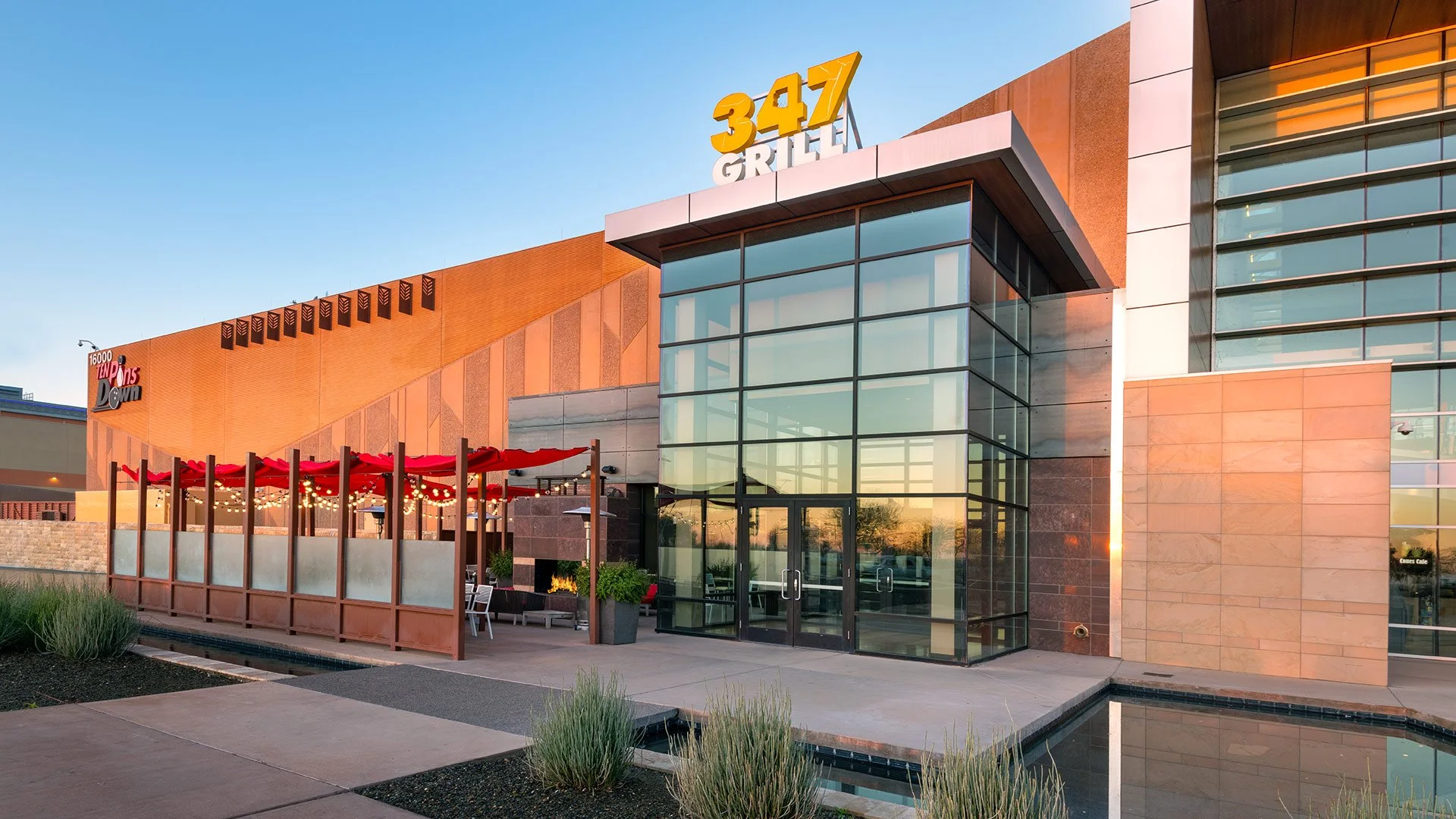 Exterior view of a modern restaurant called 347 Grill with glass entrance, outdoor seating area with red umbrellas and string lights, and a water feature in the foreground during sunset.