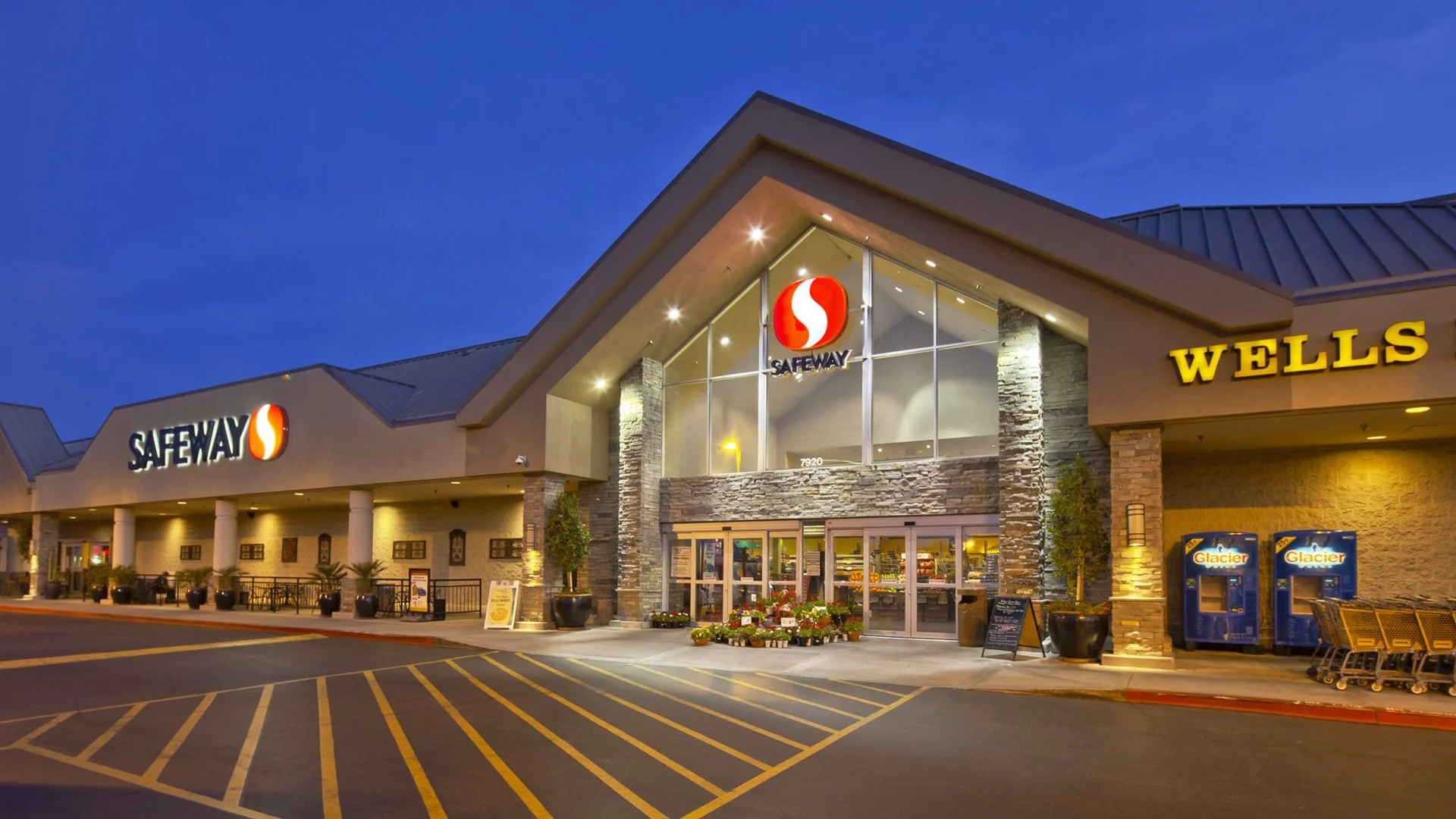 Exterior of a Safeway grocery store with a Wells sign, illuminated signs, planters, shopping carts, and self-service gas pumps, taken during twilight
