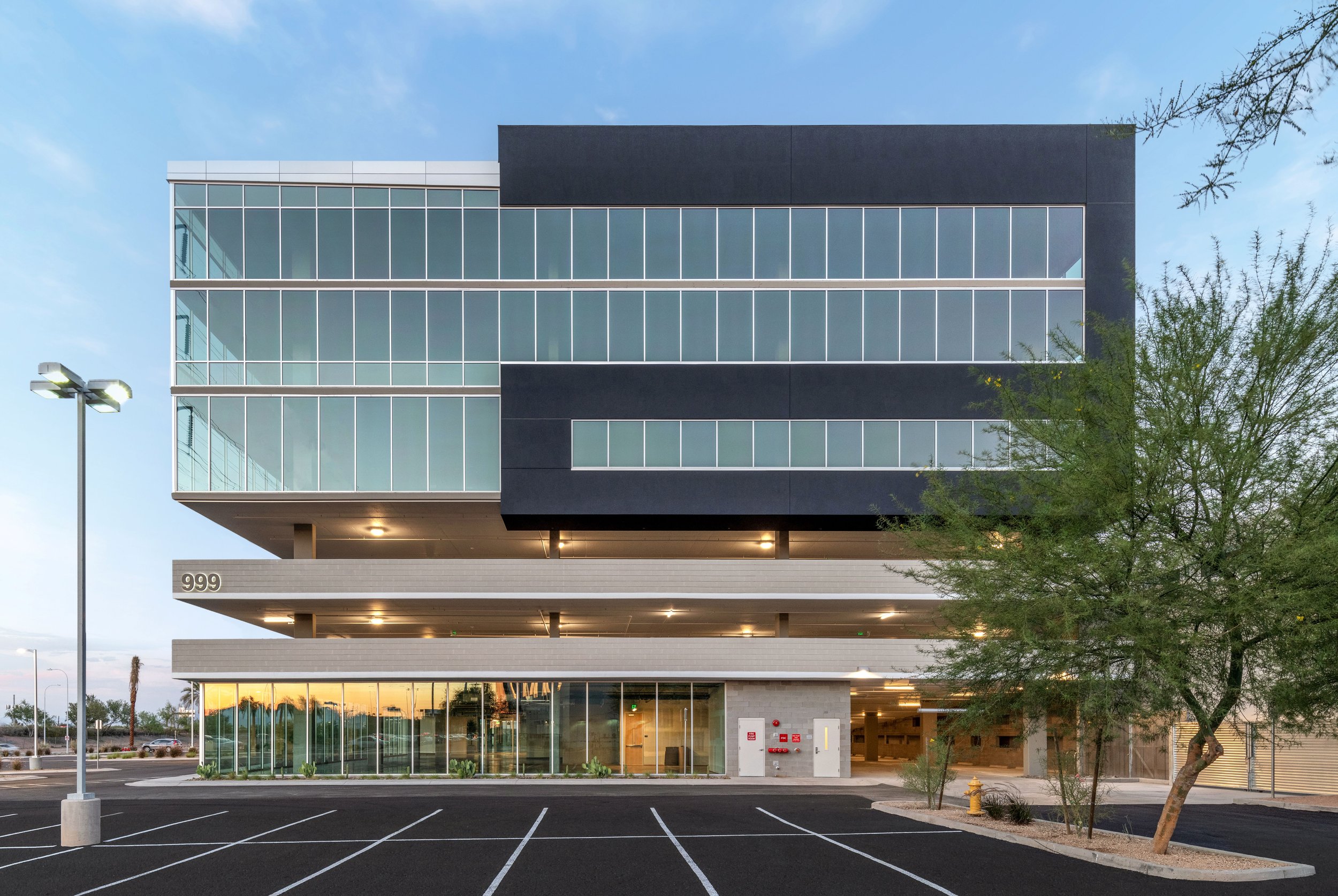 Modern multi-story office building with glass facade, covered parking below, and a parking lot in the foreground. The building has the number 999 on its lower left side.