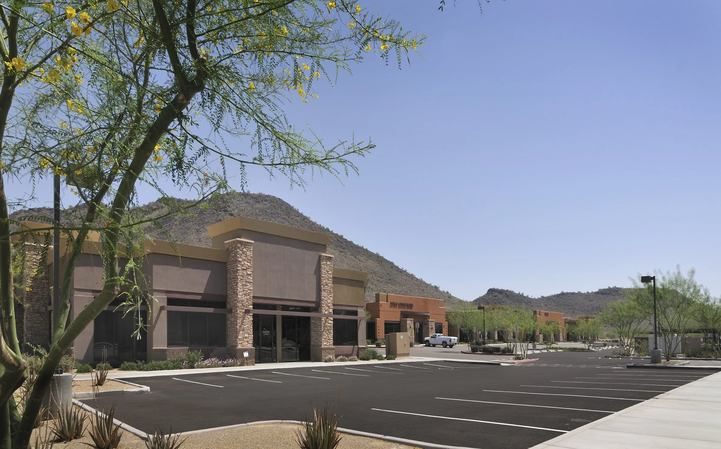An empty parking lot in front of a row of commercial buildings with a mountainous desert landscape in the background, under a clear blue sky.