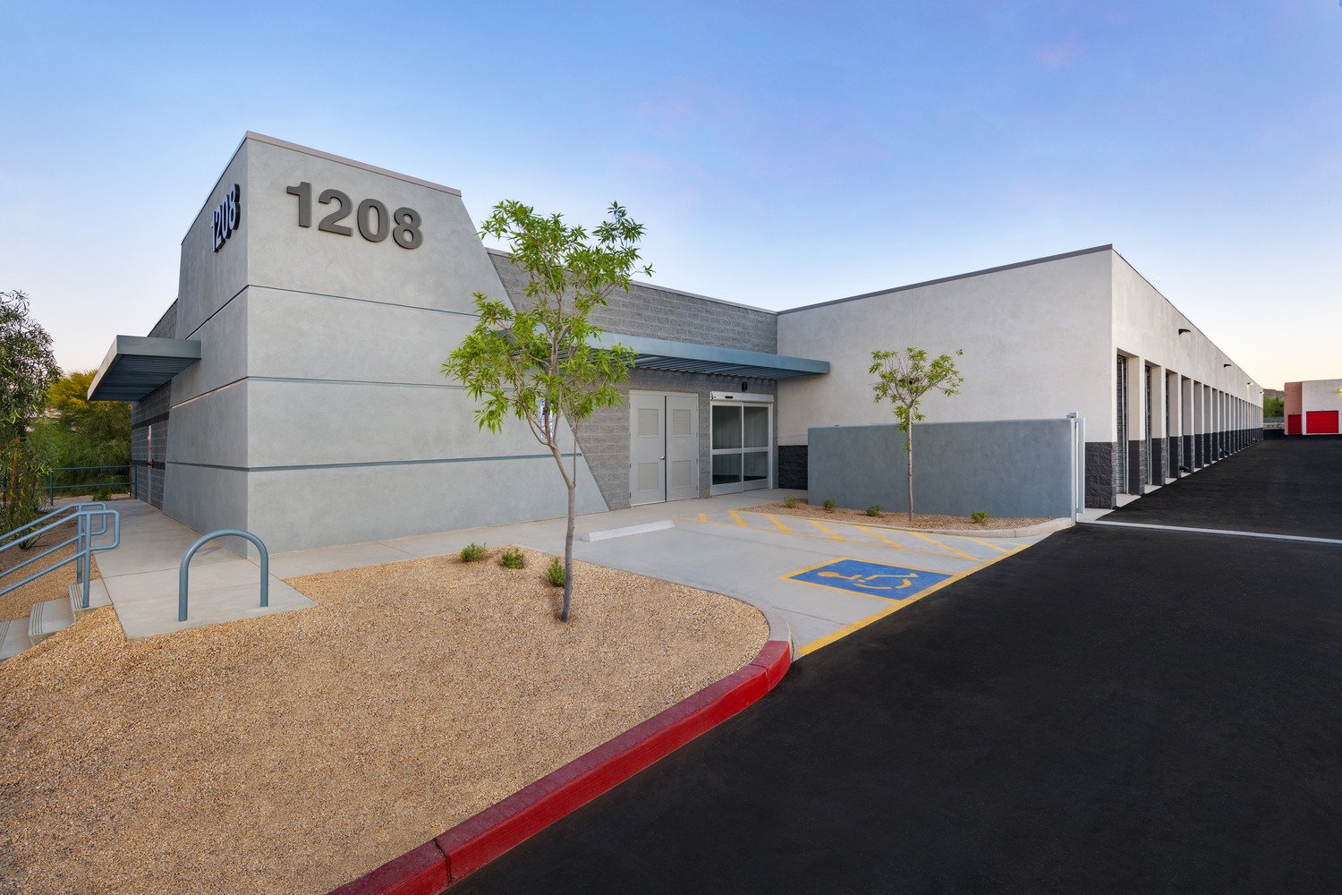 Modern commercial building with the number 1208 on its exterior wall, surrounded by trees and a parking lot with a designated handicap parking space, black asphalt, and a red painted curb.