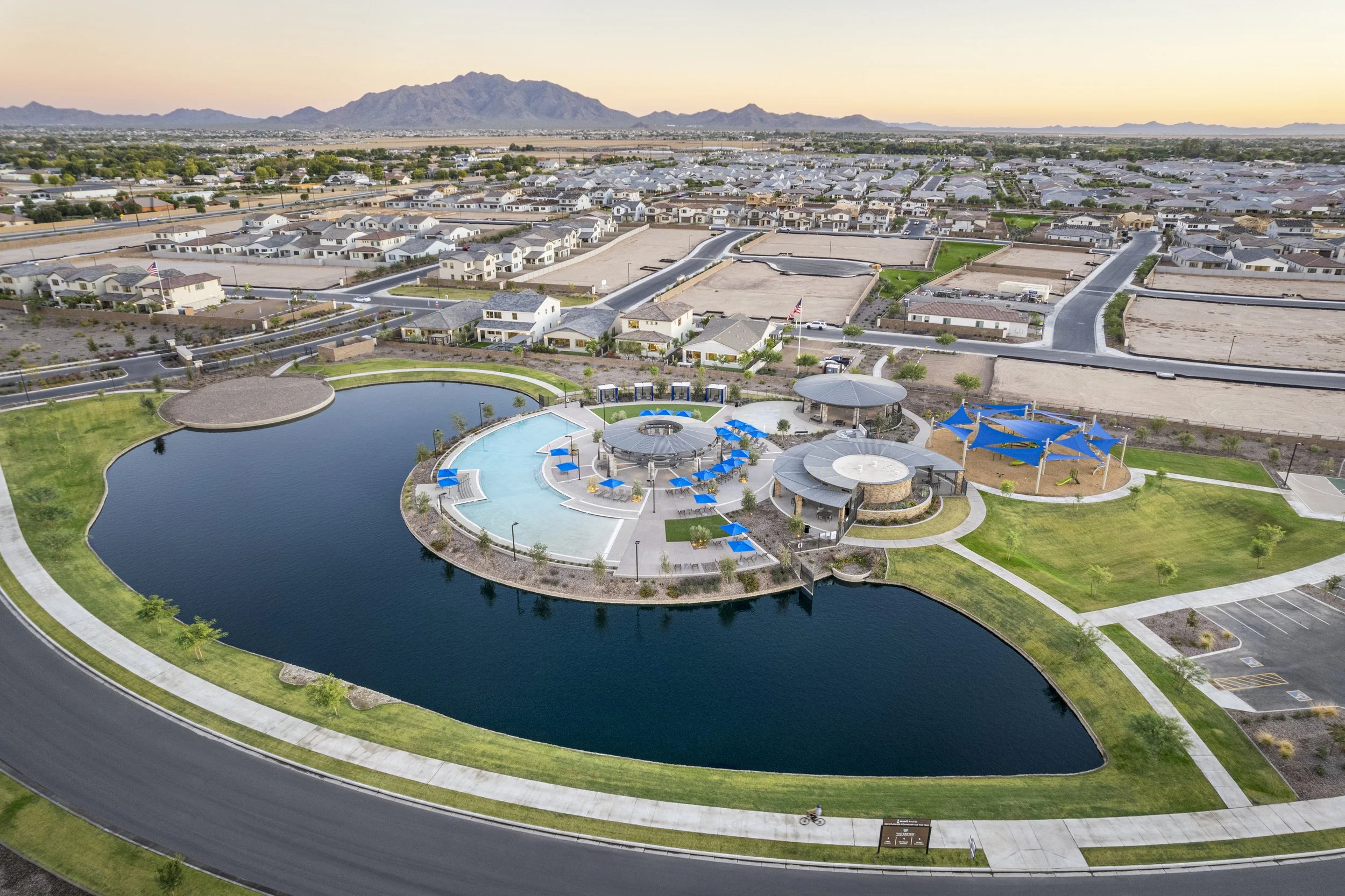 Aerial view of a community park with a large pond, swimming pool with blue chairs and umbrellas, shaded structures, green lawns, and a children's play area with blue shade sails, surrounded by residential houses and mountains in the background at sun