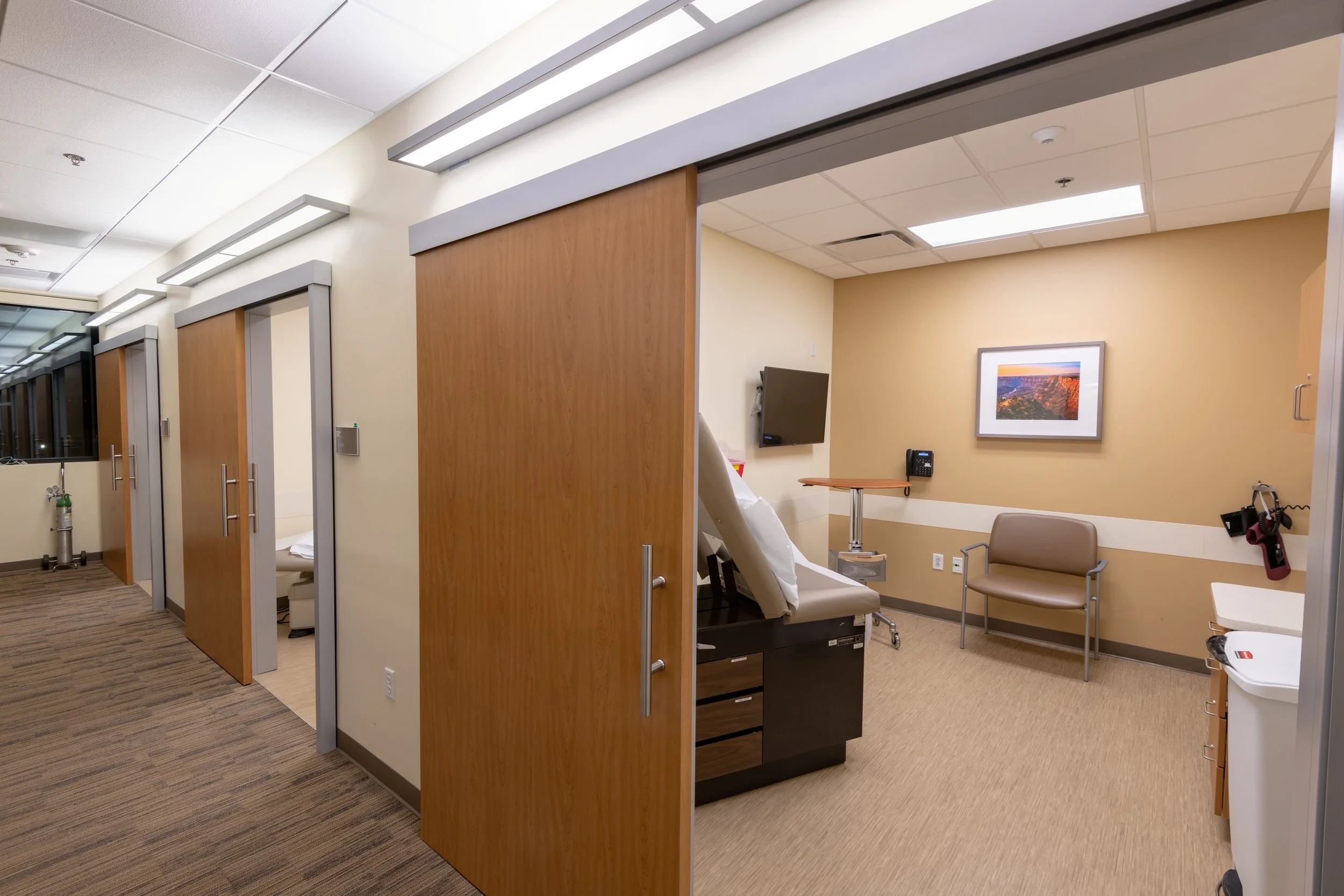 Hospital room with sliding door open showing hospital bed, television, framed artwork, and waiting chair.