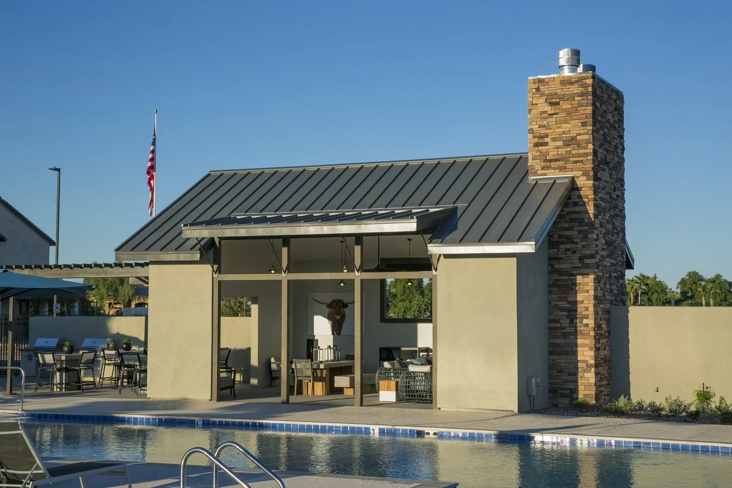 A swimming pool area with lounge chairs, a shaded seating area with a mountain goat mounted on the wall, and a building with a brick chimney, under a clear blue sky.