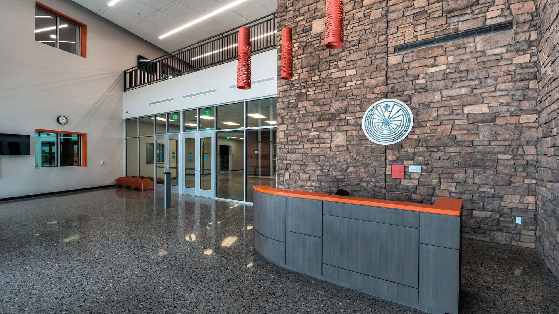 Modern lobby with a reception desk with orange accents, brick wall, decorative red hanging lights, glass doors, and a clock on the wall.