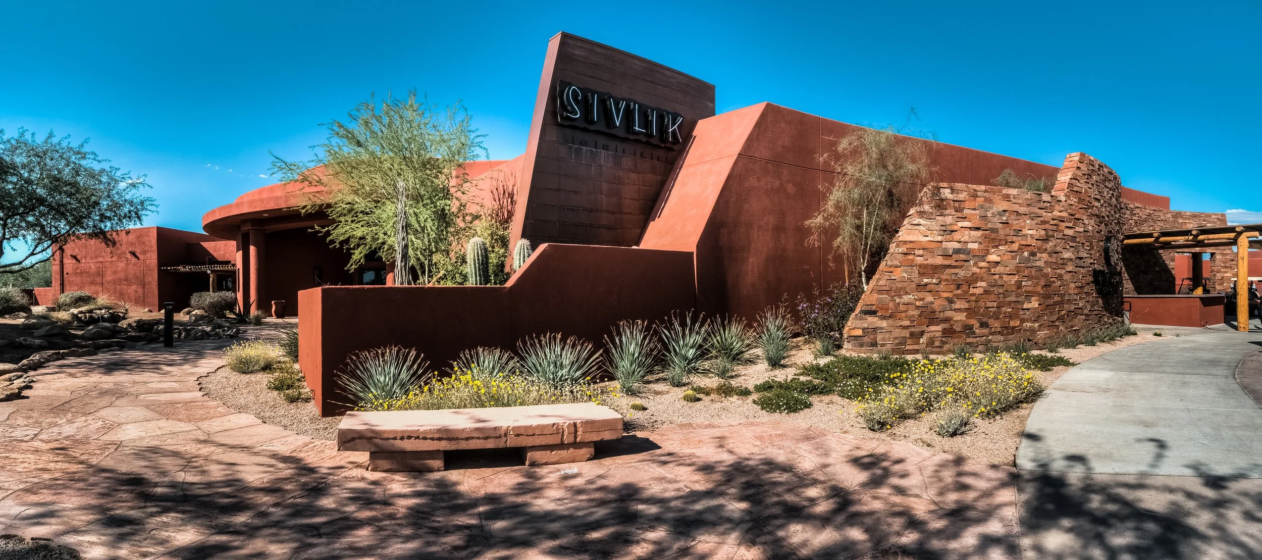 Exterior view of a modern building with a southwestern architectural style, featuring reddish-brown adobe and brick walls, desert plants, and a clear blue sky.