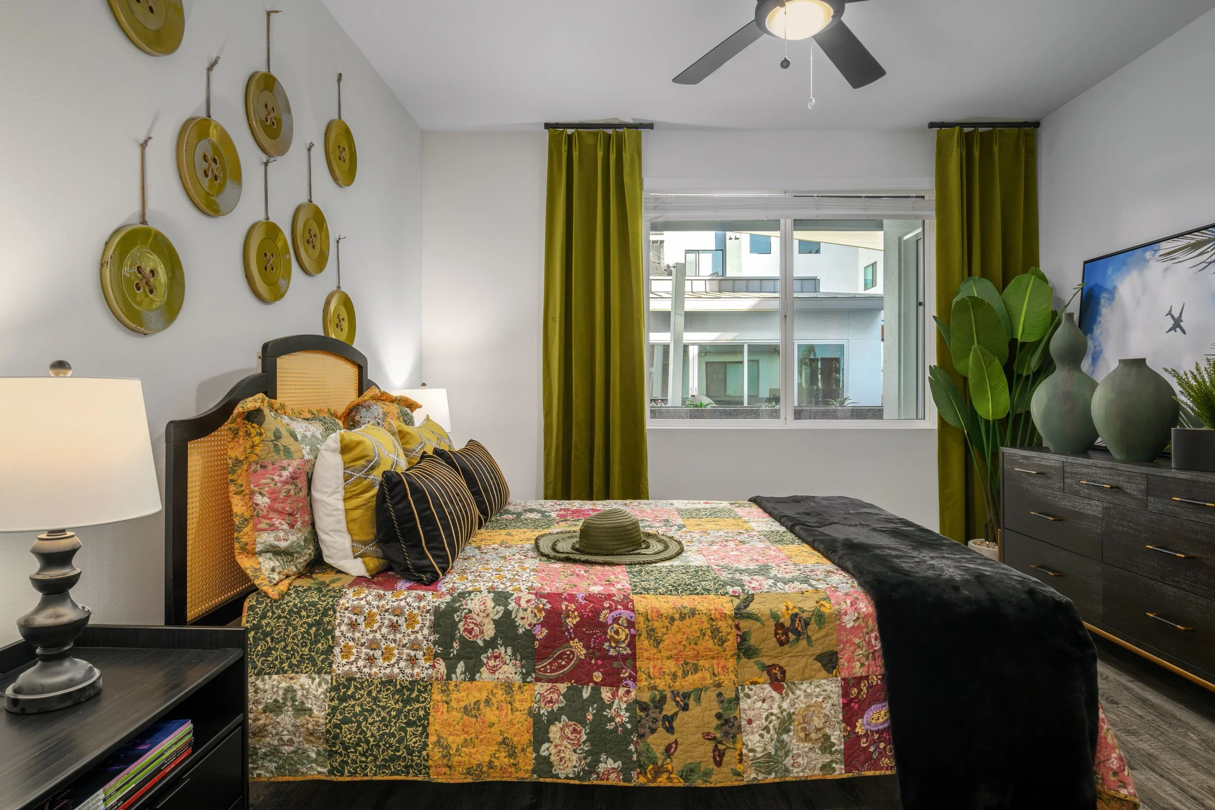 A bedroom with a colorful patchwork quilt on the bed, black pillows with gold stripes, and a straw hat. The room has white walls, green curtains, a large window, a black dresser with large vases, a small black side table with a lamp, decorative yello