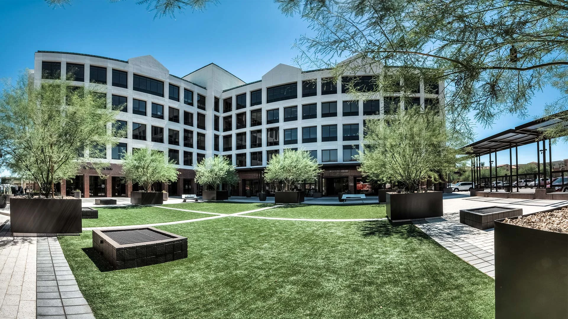 Modern multi-story office building surrounding a landscaped courtyard with trees, benches, and paved walkways under a clear blue sky.