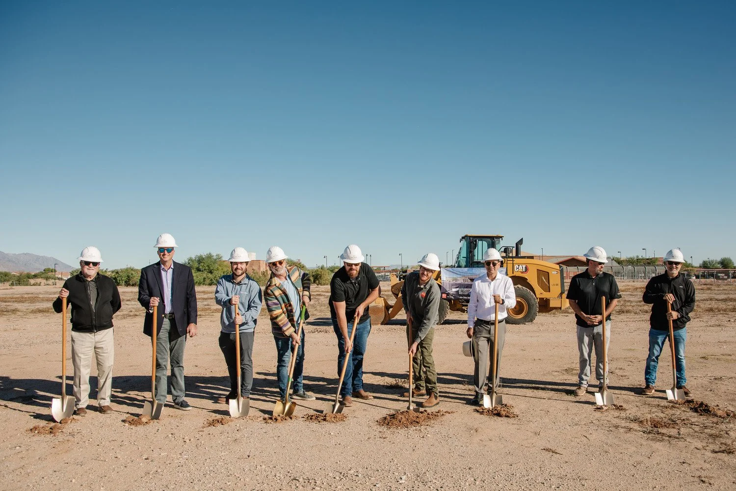 Group of ten people in suits and casual clothes, wearing white construction helmets, holding shovels, standing in a line on a construction site with dirt ground, a yellow bulldozer in the background, and clear blue sky.