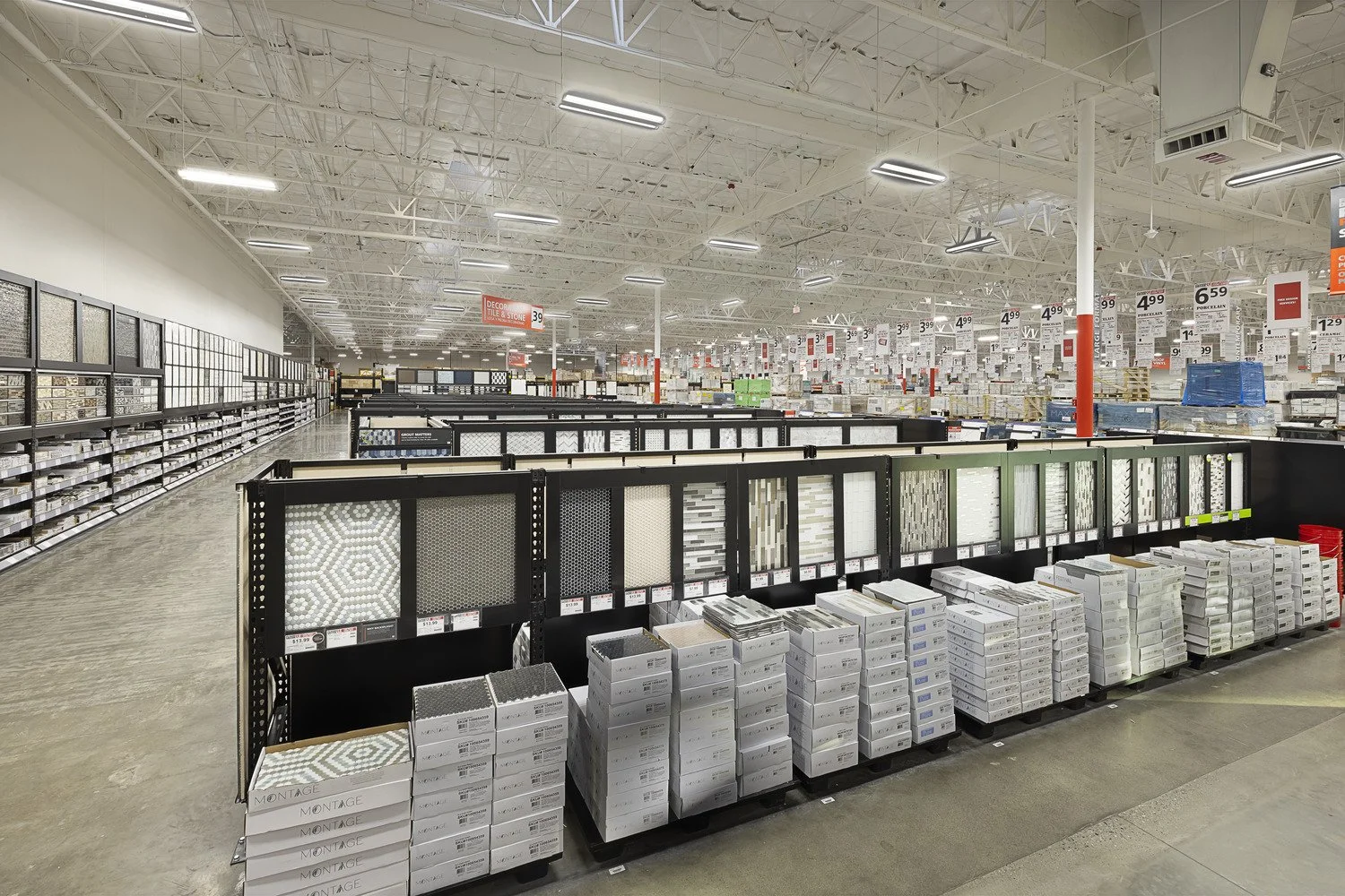 Empty store aisle with tile samples displayed on black tiered racks in a large retail store, fluorescent lights hanging from a white ceiling, and price signs overhead.