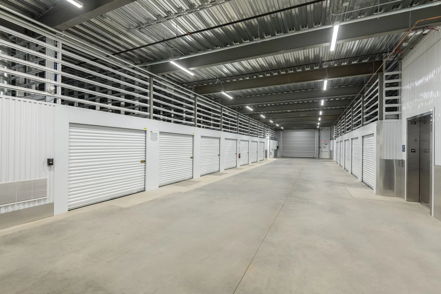 Empty indoor storage unit facility with white rolling doors, concrete floor, and metal ceiling with lighting.