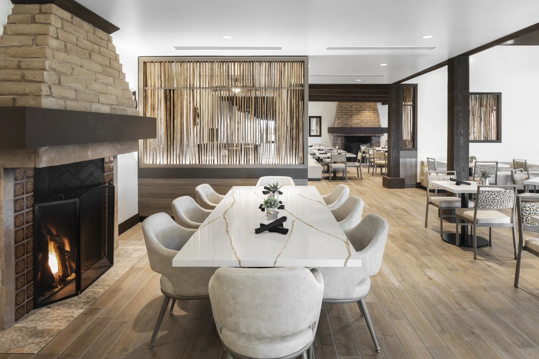 Modern restaurant interior with a long marble dining table surrounded by beige upholstered chairs, brick fireplace on the left, wooden flooring, and a decorative wooden partition.