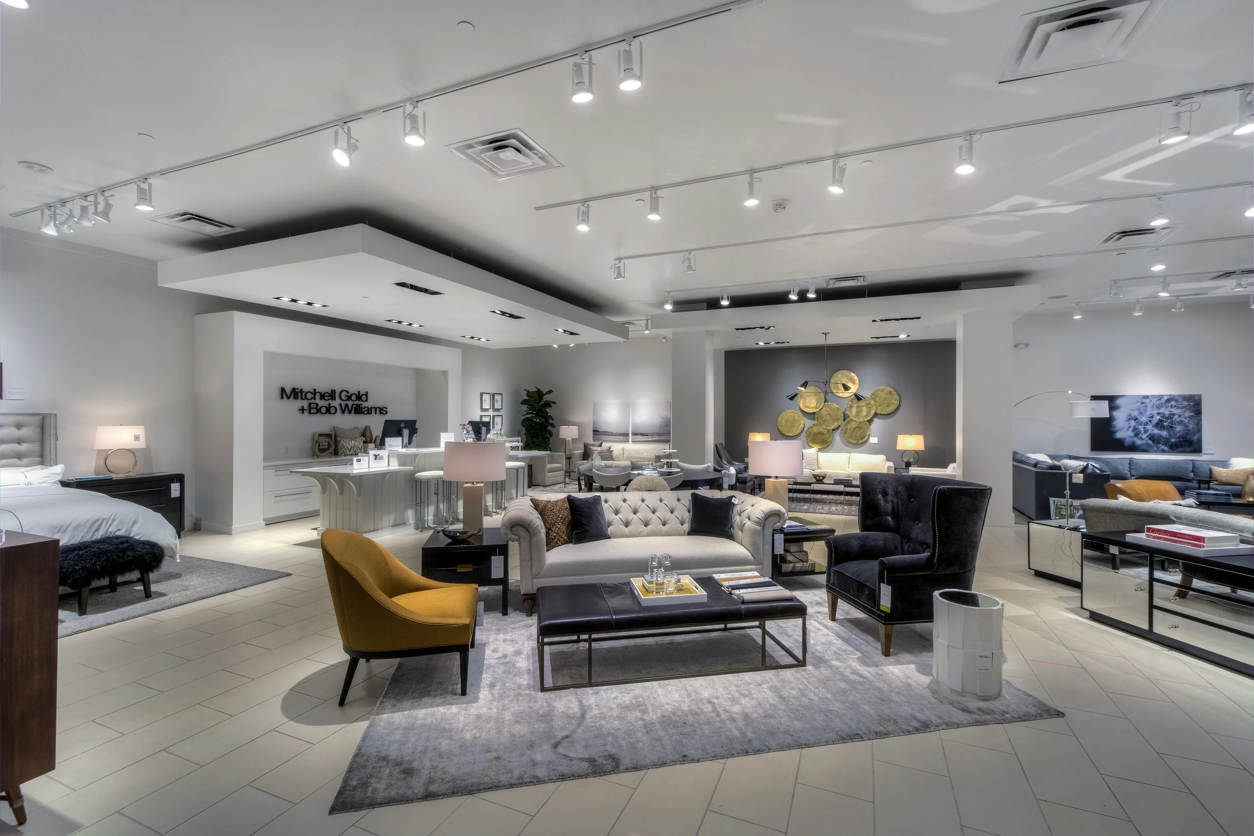 Modern furniture showroom with sofas, armchairs, coffee tables, lamps, and decorative wall art in a clean, well-lit space.