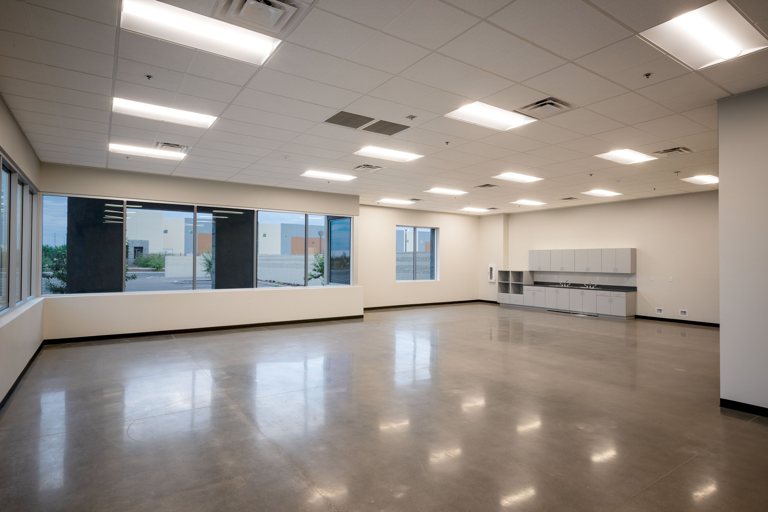 Empty commercial room with large windows, fluorescent lighting, and kitchenette in the corner.
