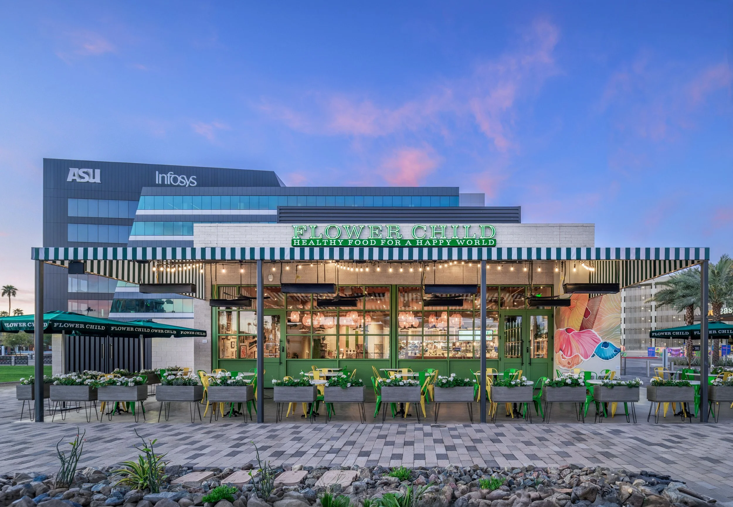 Exterior of Flower Child restaurant with outdoor seating, green chairs, white planters, striped awning, and Cityscape background with ASU and Infosys buildings, during sunset.