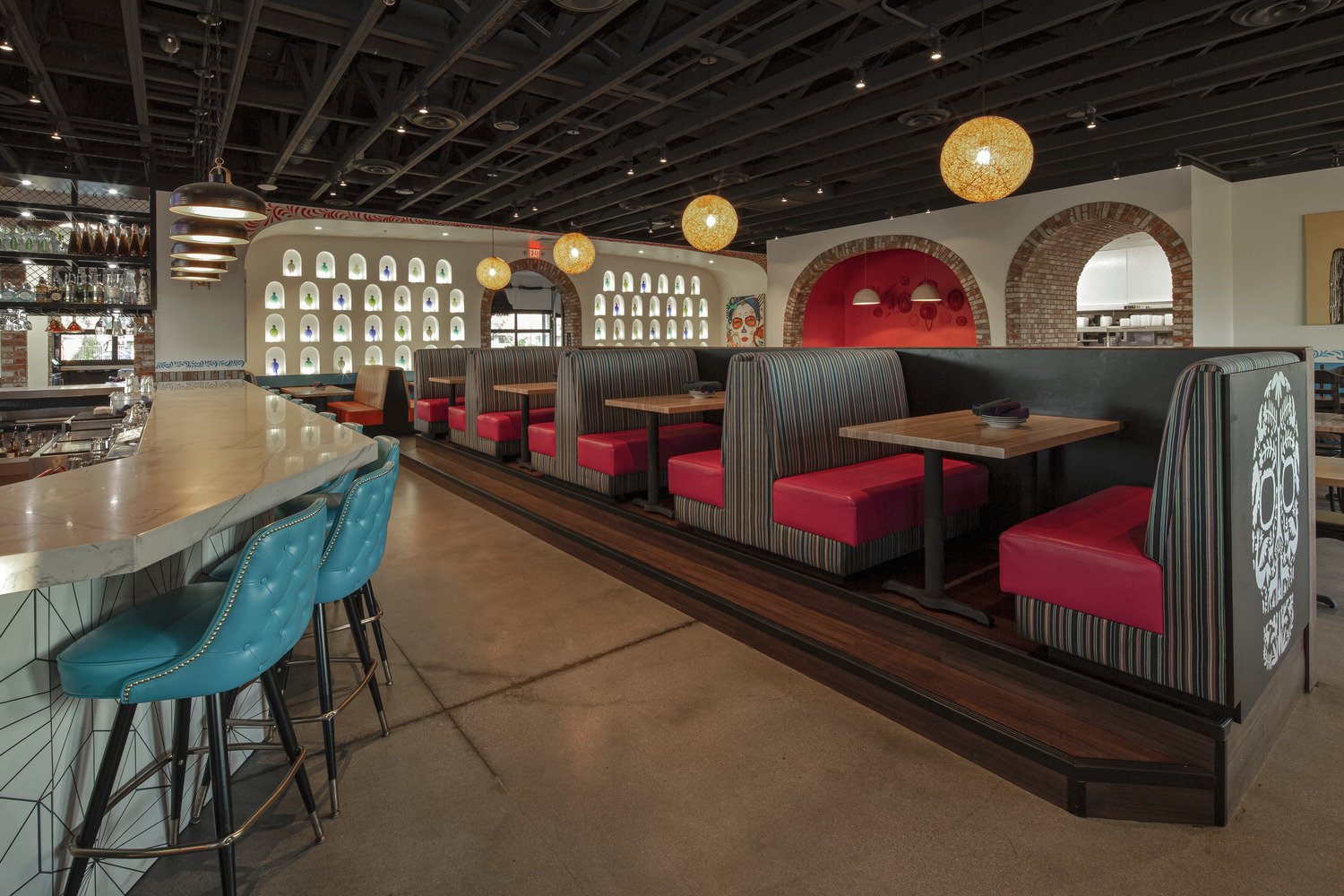 Interior of a restaurant with a bar area, turquoise barstools, booth seating with red cushions, decorative wall niches with colorful objects, and warm lighting.