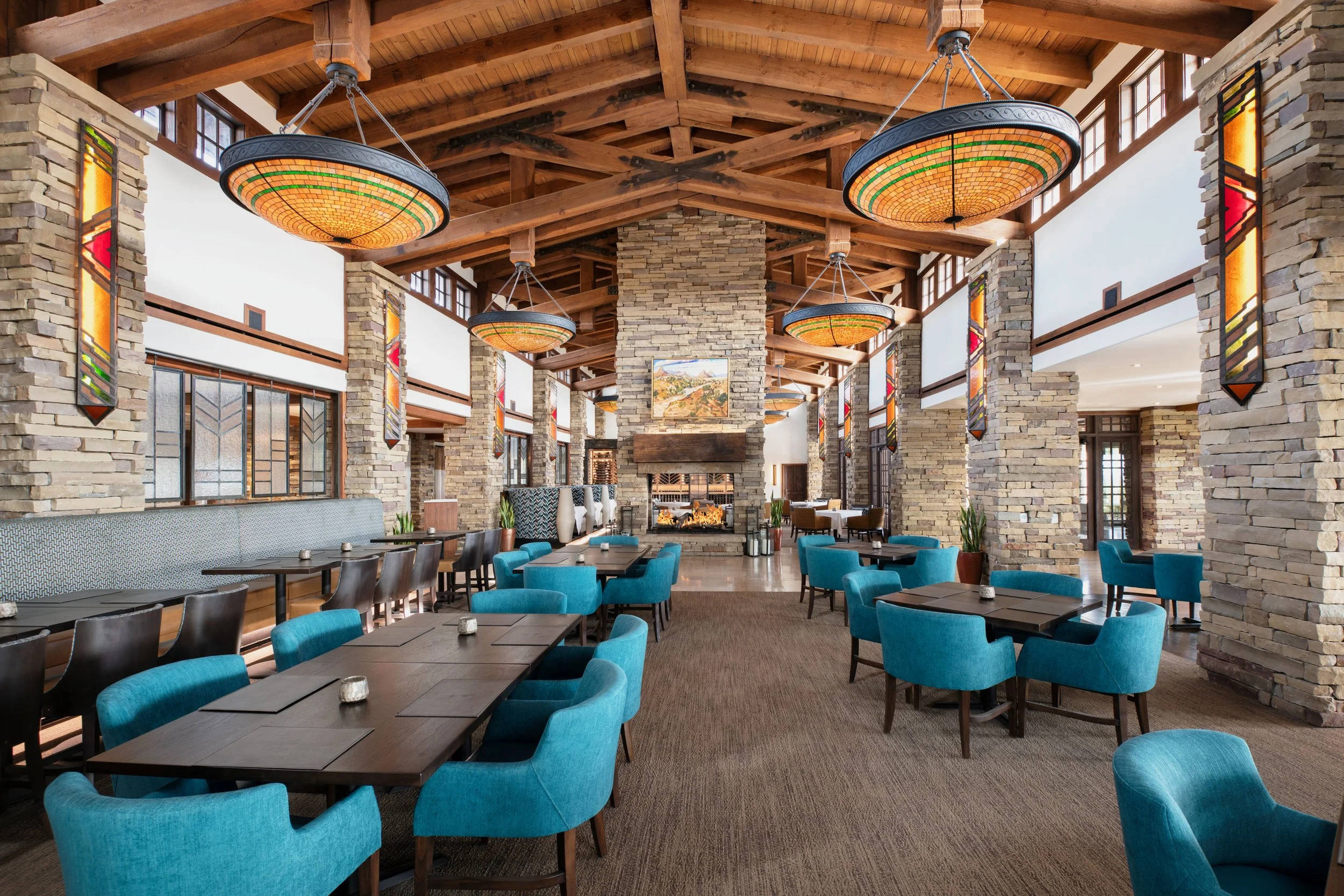 Interior of a restaurant or lounge with stone walls, wooden beams, blue cushioned chairs, dark wood tables, stained glass lighting fixtures, and a fireplace in the background.