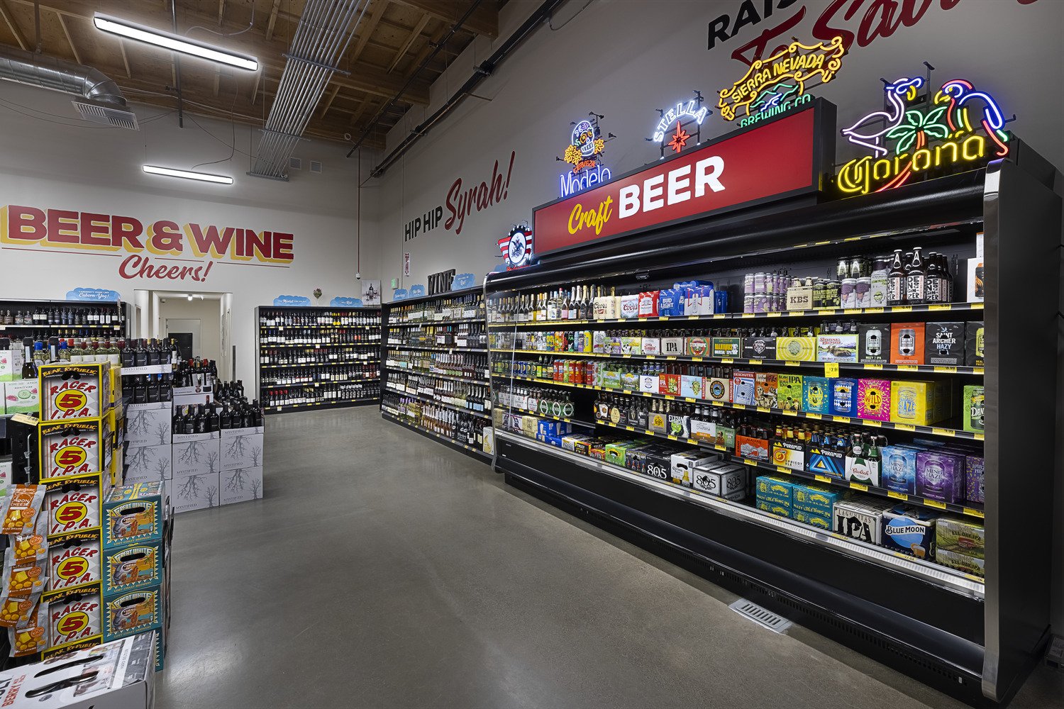 Interior of a store aisle with shelves stocked with craft beer and alcoholic beverages, neon signs with beer brands, and signs that read 'Beer & Wine Cheers!' and 'Hip Hip Syrah!' on the wall.