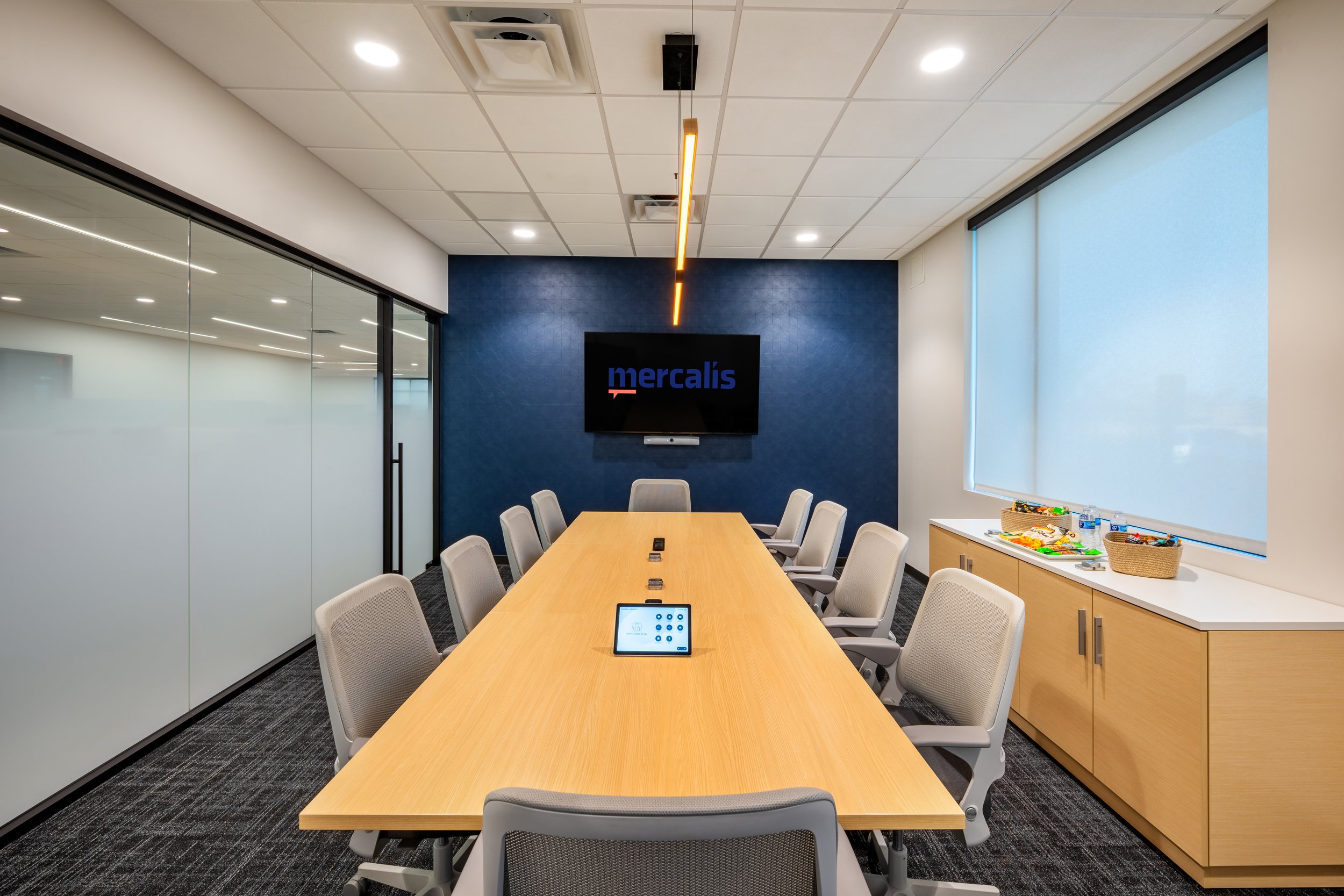 Conference room with a long wooden table, beige chairs, a wall-mounted TV screen displaying the word 'mercalis,' a tablet on the table, a blue accent wall, large windows with blinds, and a side cabinet with snacks and water bottles.