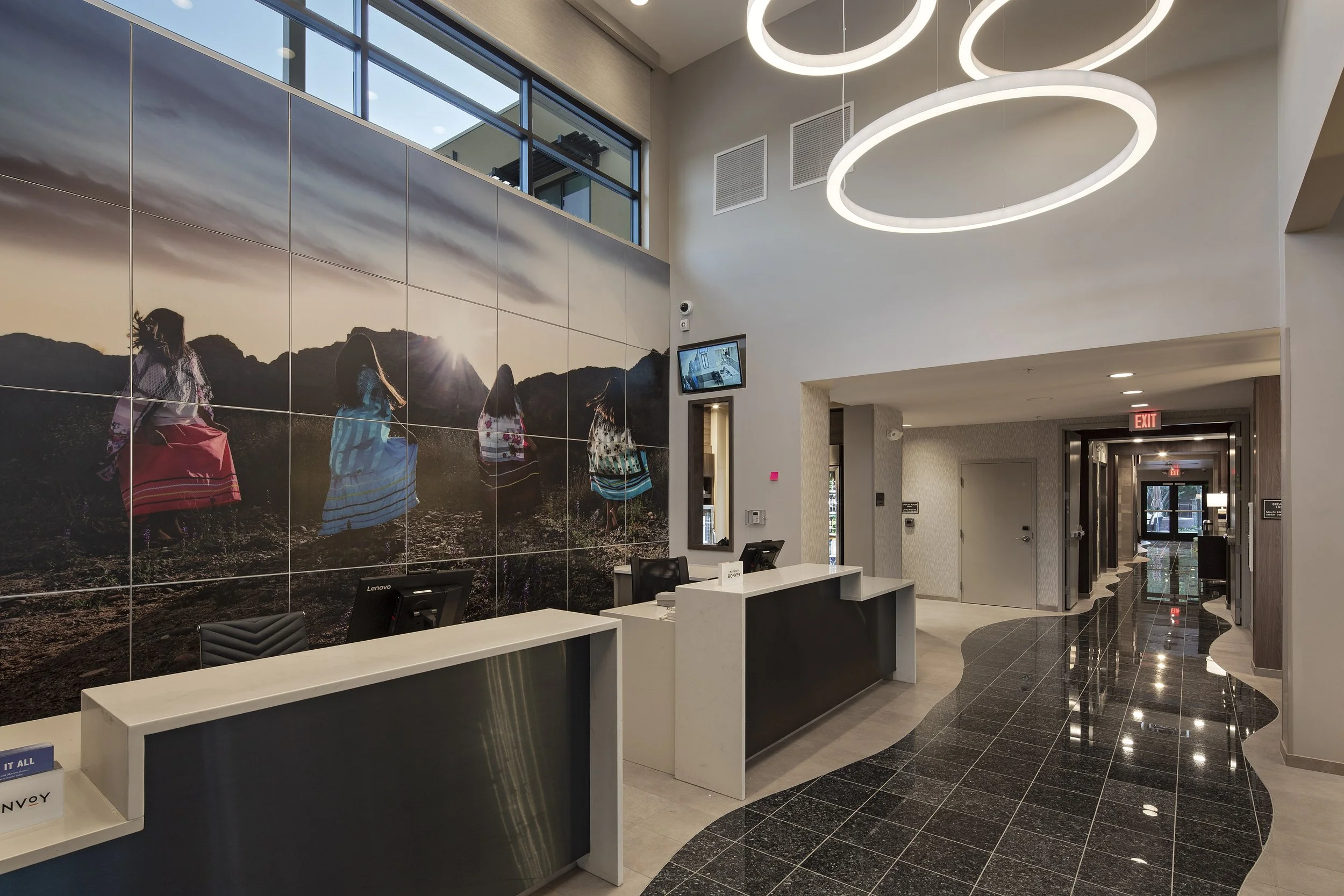 Hotel lobby with front desk, large mural of women in colorful dresses on mountains, modern lighting fixtures, and hallway with glossy black and beige flooring.