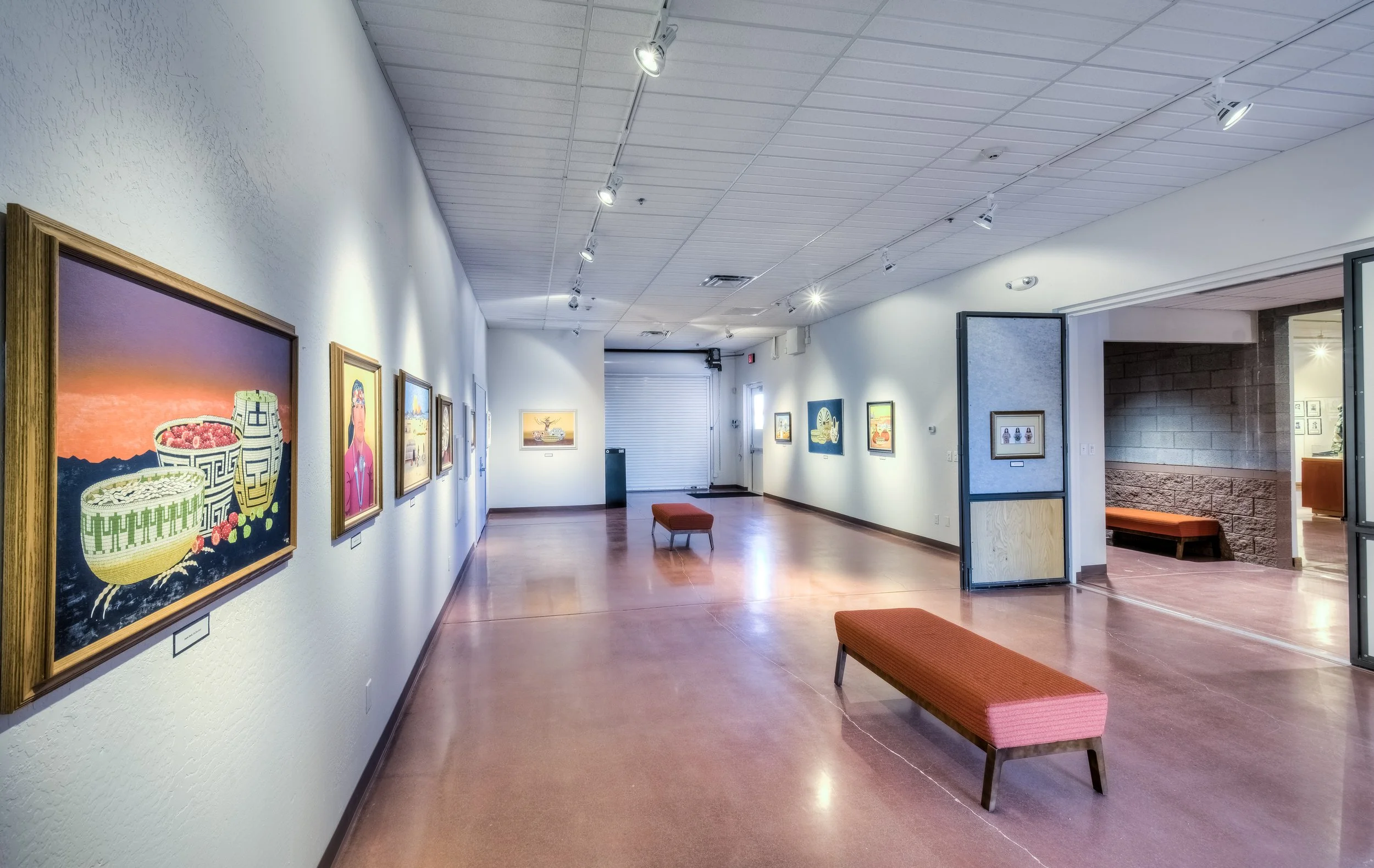 Art gallery with colorful paintings on white walls, wooden benches, and track lighting.